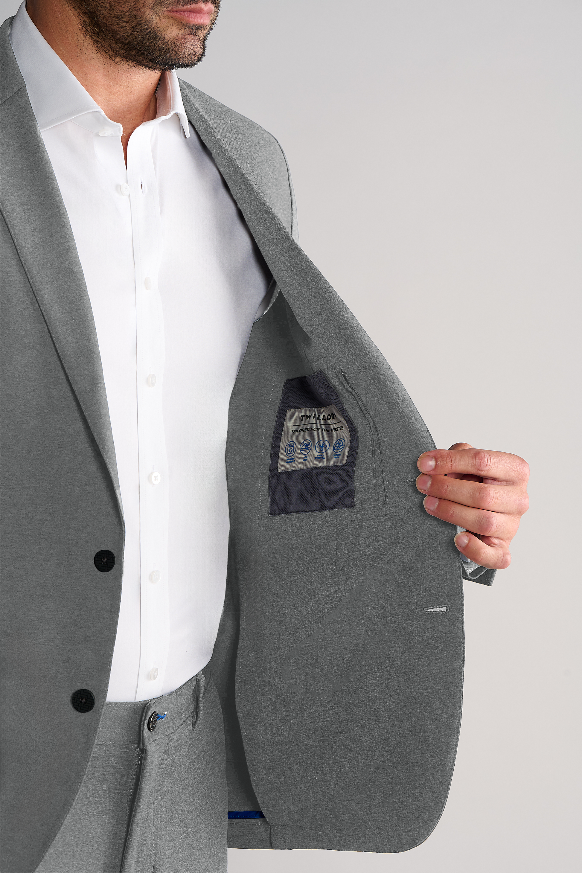 A man in a Twillory Performance Blazer // Heather Light Grey and white shirt holds open his jacket to reveal the inside pocket and label. His face is partially out of frame against a plain light grey background.