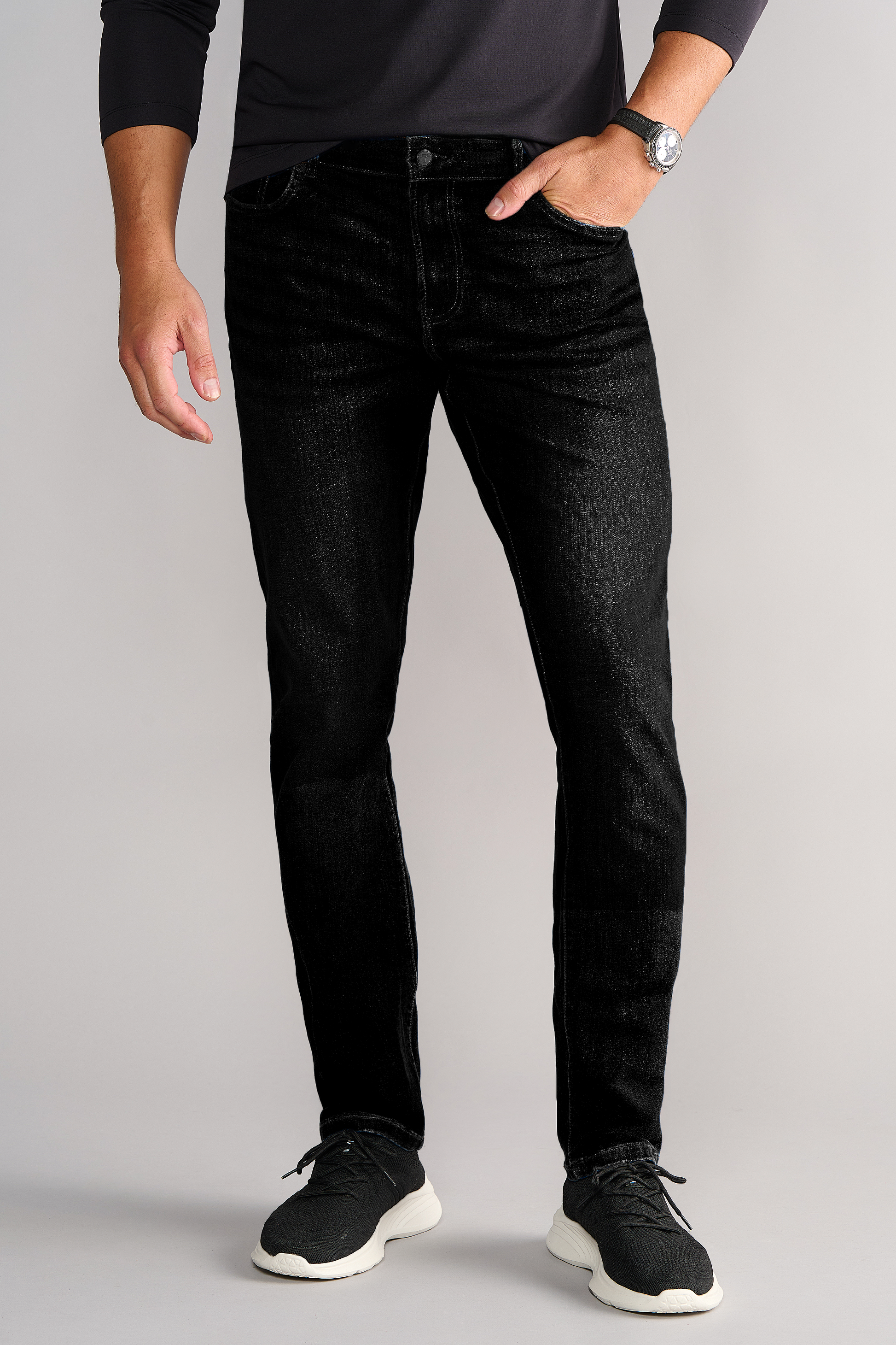 A man wearing Twillory's Performance Denim // PURE BLACK jeans, a black long-sleeve shirt, a silver watch, and black sneakers stands against a light gray background. Only his body from the shoulders down is visible; left hand in pocket.
