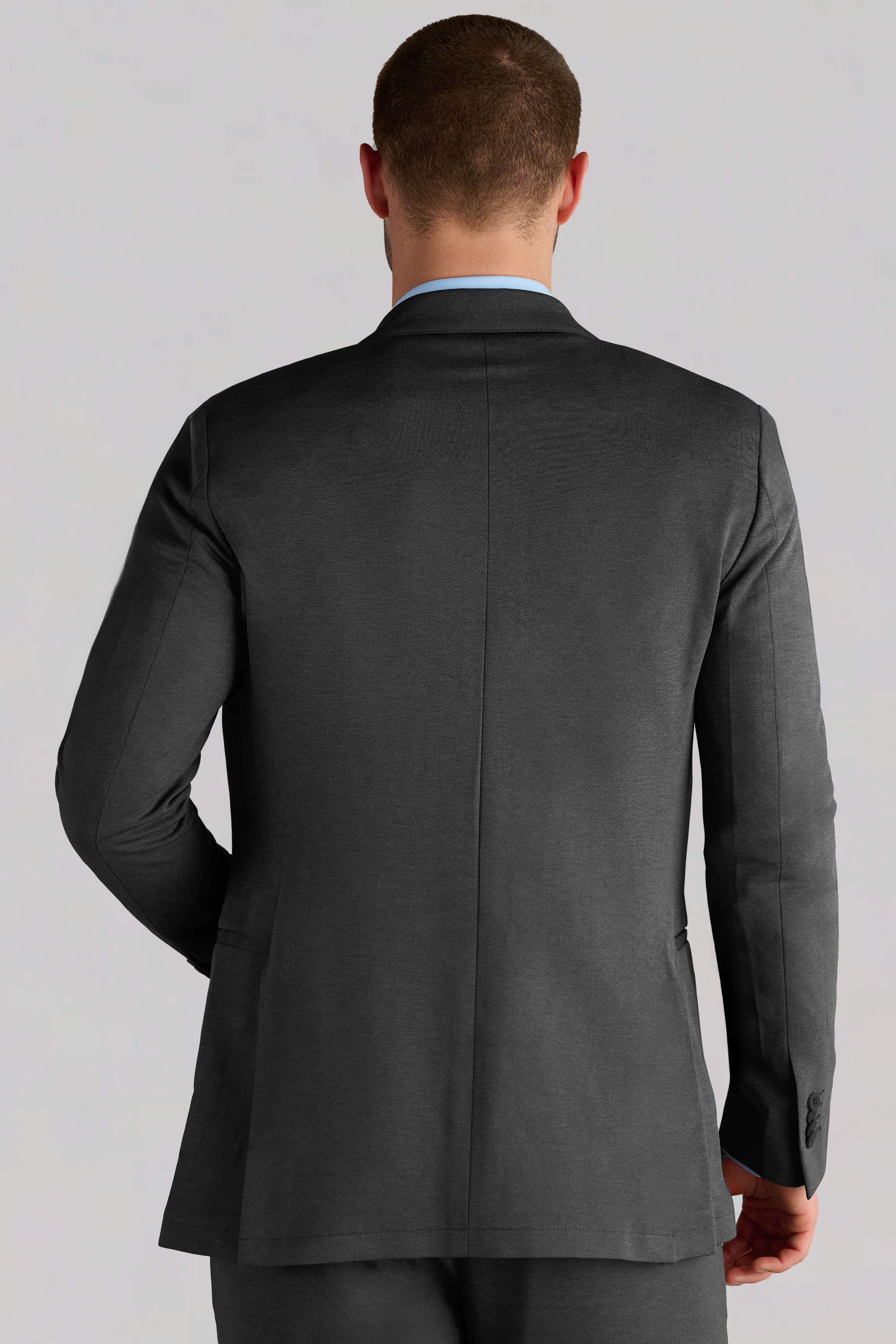 A man with short hair, seen from behind, wears the Twillory Performance Blazer // Charcoal with matching pants. His left arm is slightly bent, showing subtle seams and cuff buttons. The background is light and plain.