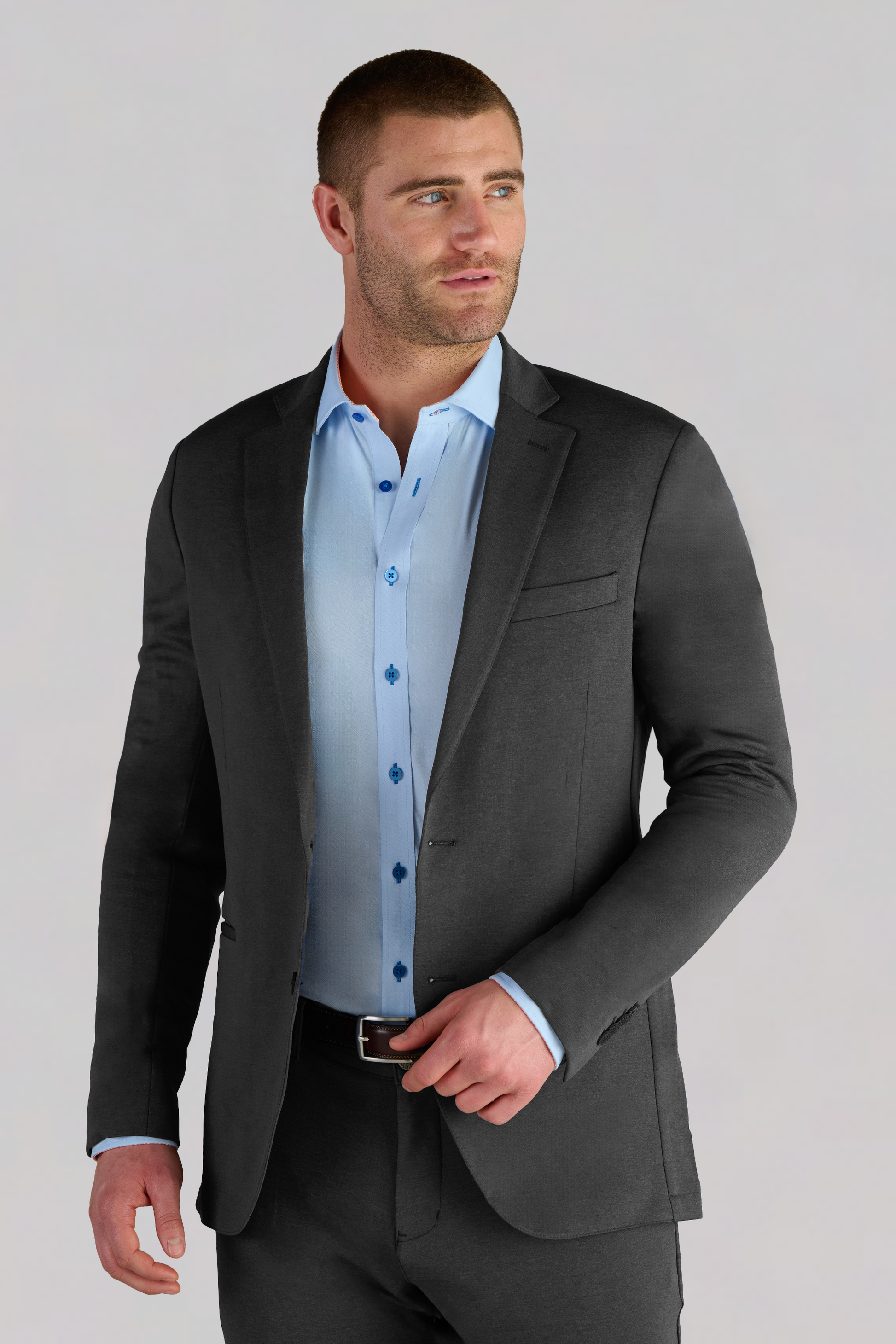 A man with short brown hair and a trimmed beard wears the Twillory Performance Blazer in Charcoal over a light blue dress shirt. He stands against a plain light gray background, facing slightly right and adjusting his belt.