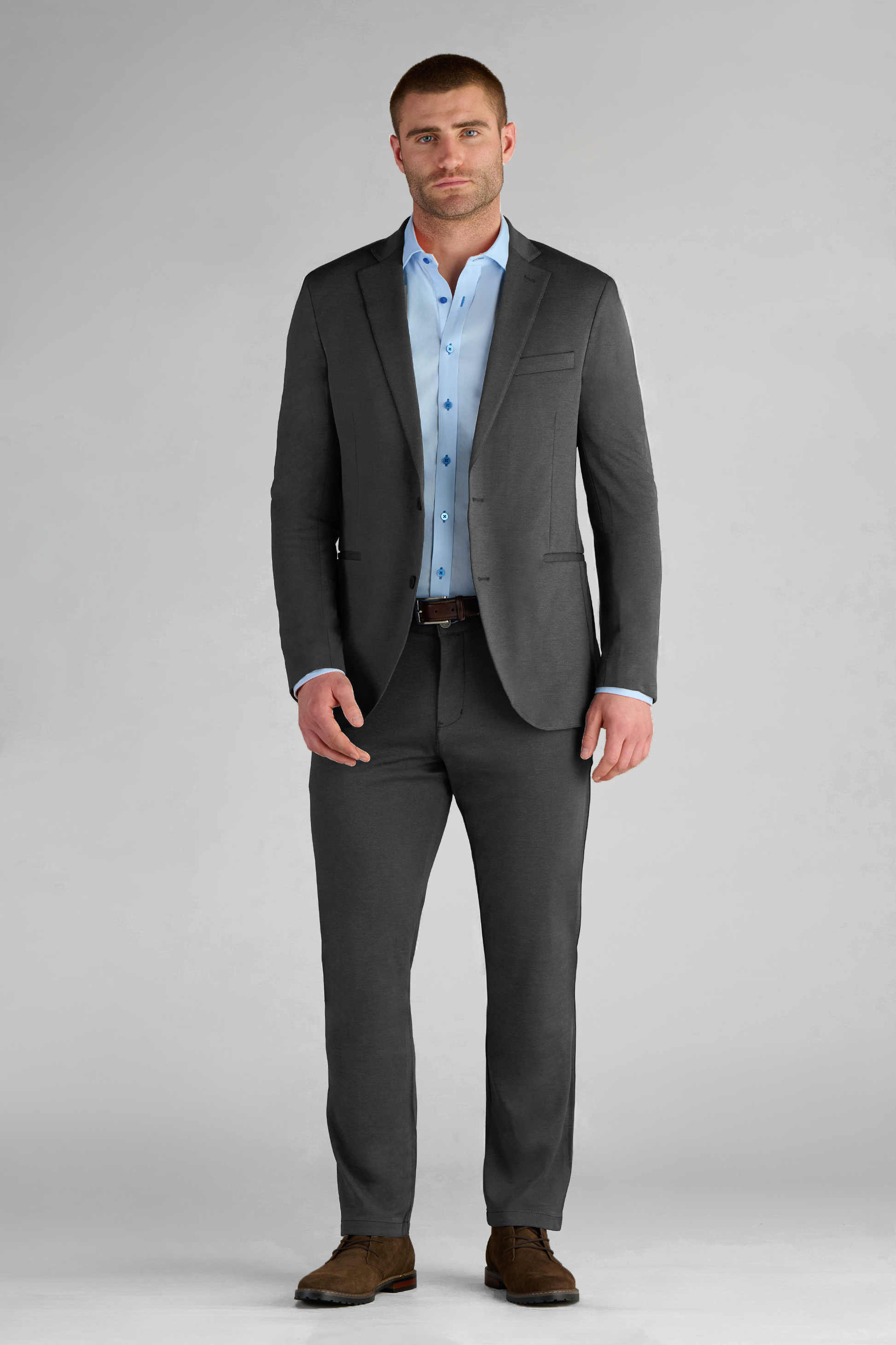 A man with short hair and a trimmed beard stands facing forward against a light gray background, wearing Twillory’s Performance Blazer in Charcoal, paired with a light blue dress shirt and brown dress shoes.