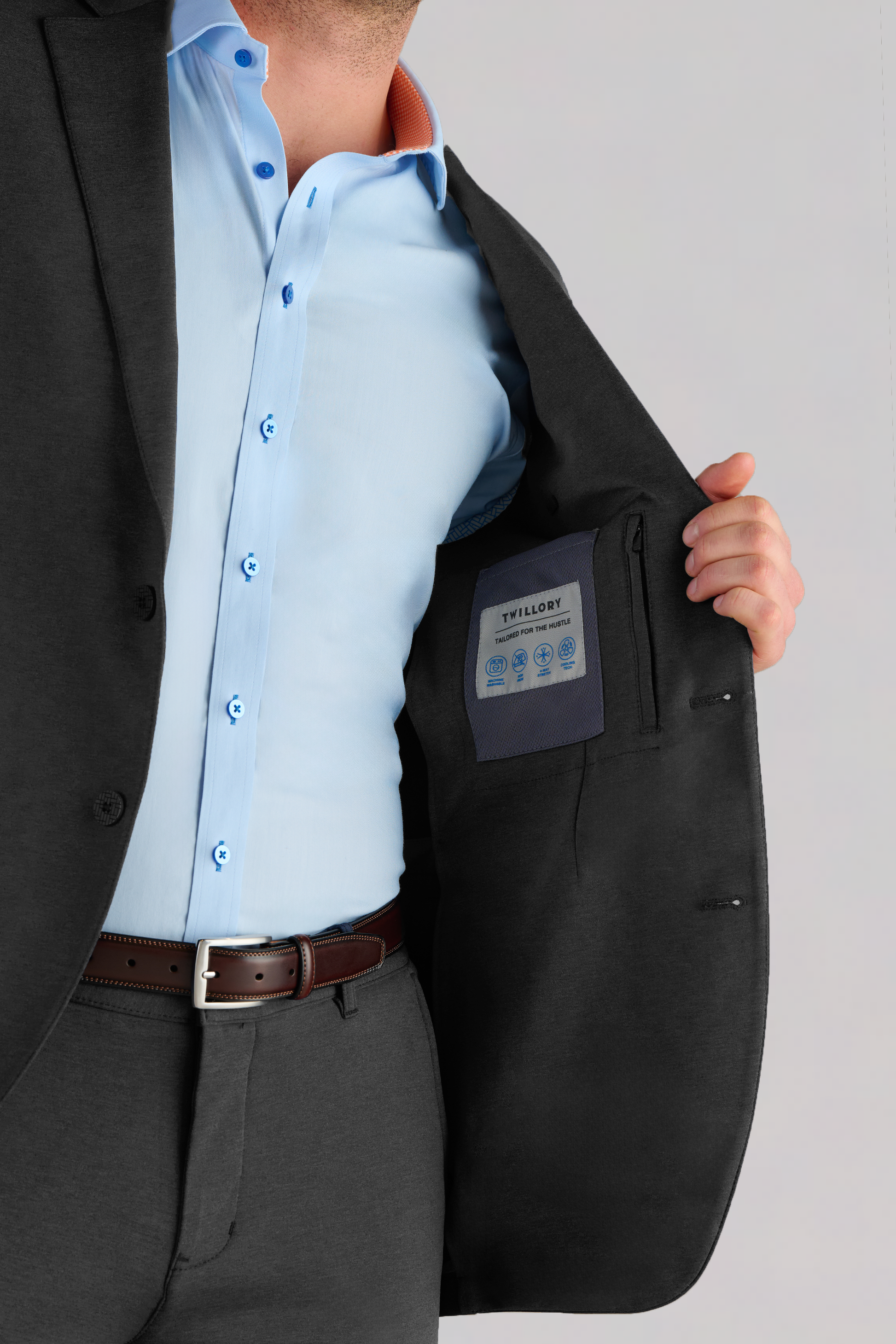 A man in a dark suit and light blue shirt opens his Twillory Performance Blazer // Charcoal to reveal the inner pocket with branded icons and text. His face is not visible as he holds the jacket open with his right hand.