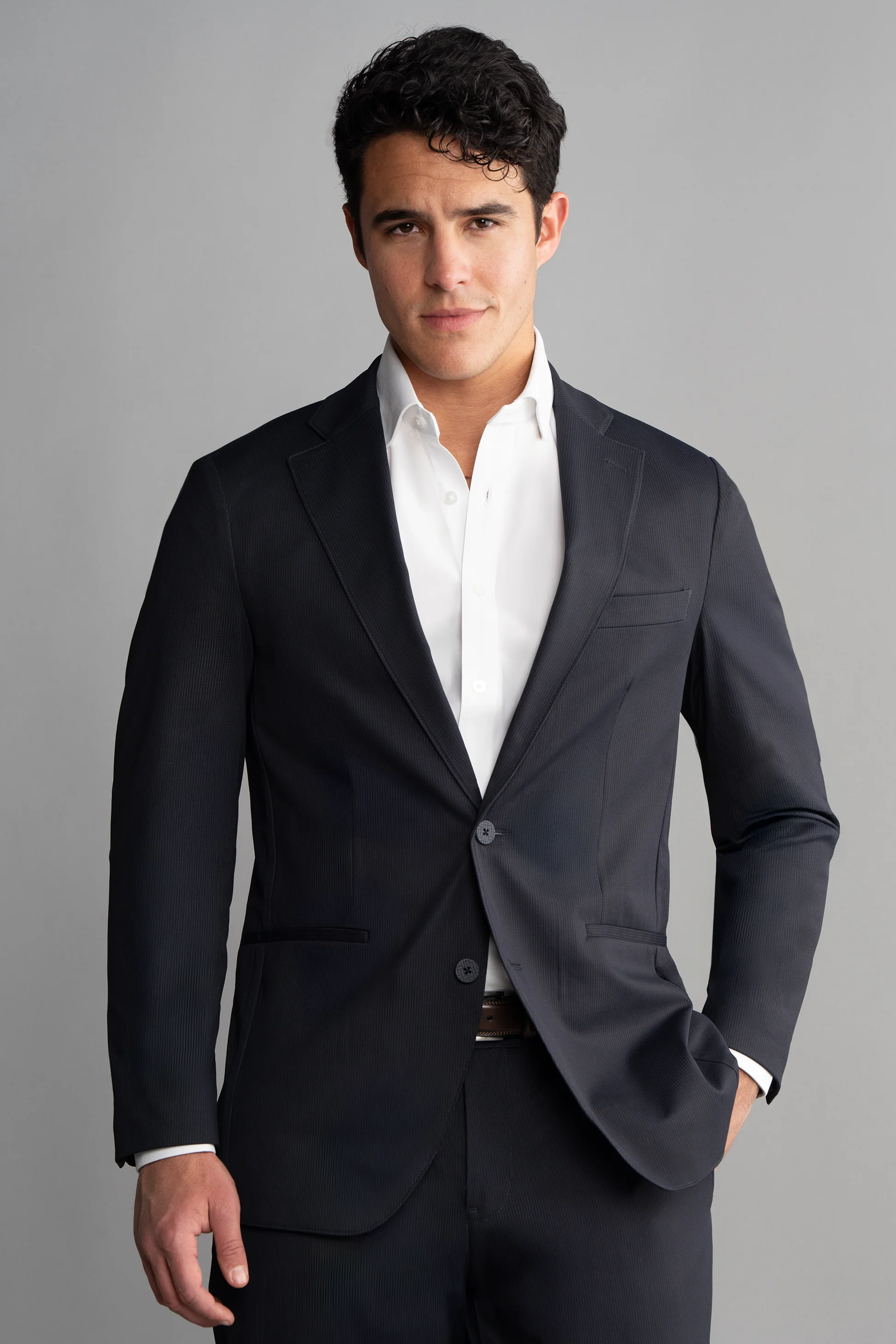 A man with short, dark curly hair wears the Twillory Performance Blazer in Navy Pin Stripe over a white dress shirt. He stands against a plain gray background, looking confidently at the camera with one hand in his pocket.