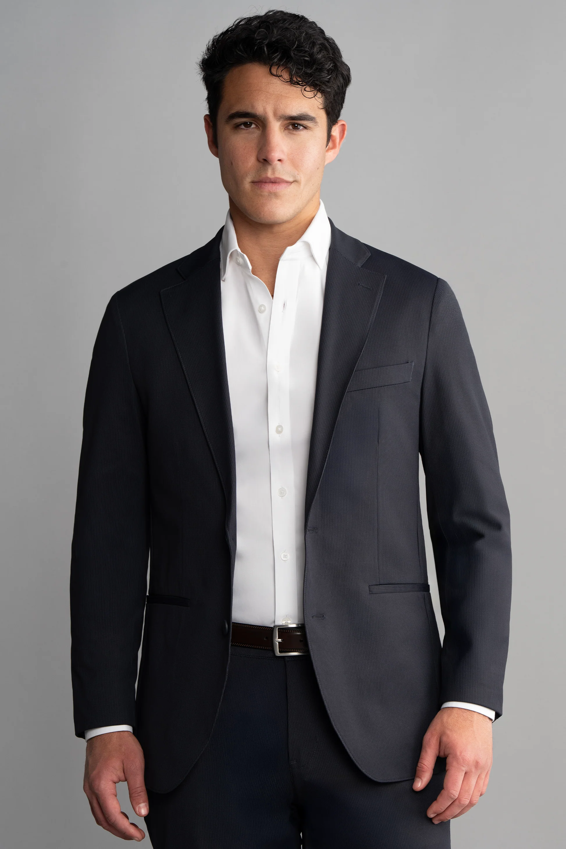A man with short dark hair wears the Twillory Performance Blazer in Navy Pin Stripe over an unbuttoned white shirt. He stands facing forward against a light gray background, arms relaxed and expression neutral.