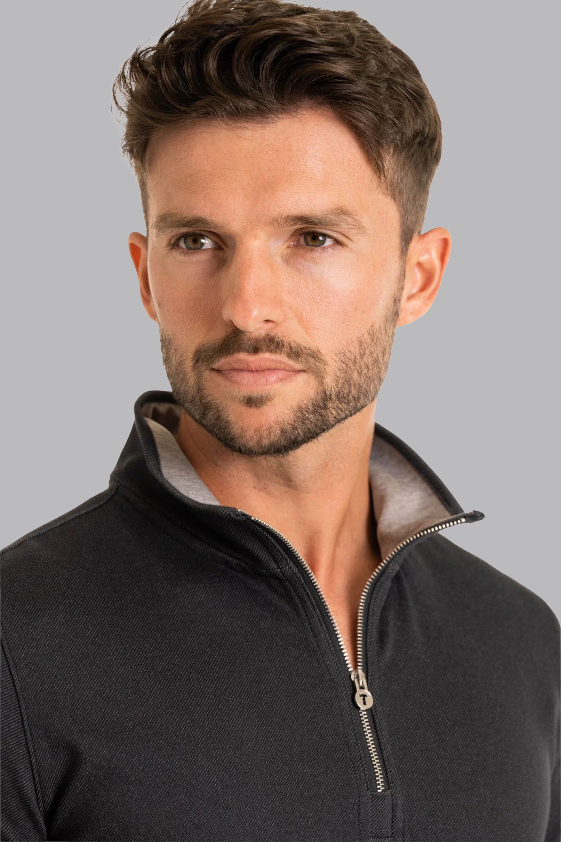 A man with short brown hair and a trimmed beard looks to the side in a Twillory Performance Quarter Zip in Charcoal, featuring a light gray inner collar. He has a neutral, contemplative expression against a plain light gray background.