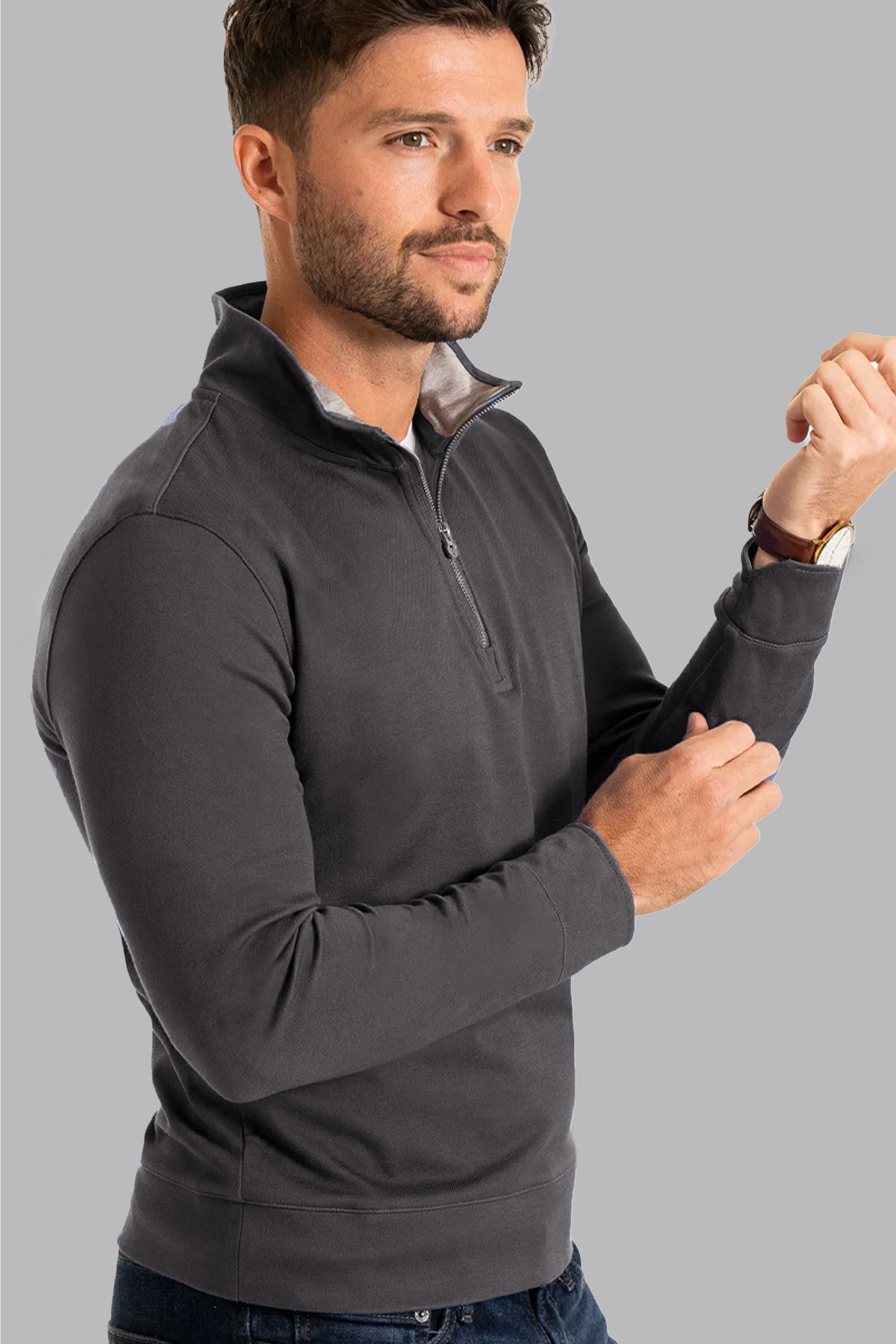 A man with short brown hair and a beard models the Twillory Performance Quarter Zip in Charcoal, adjusting his left sleeve and wearing a watch, against a plain light gray background.