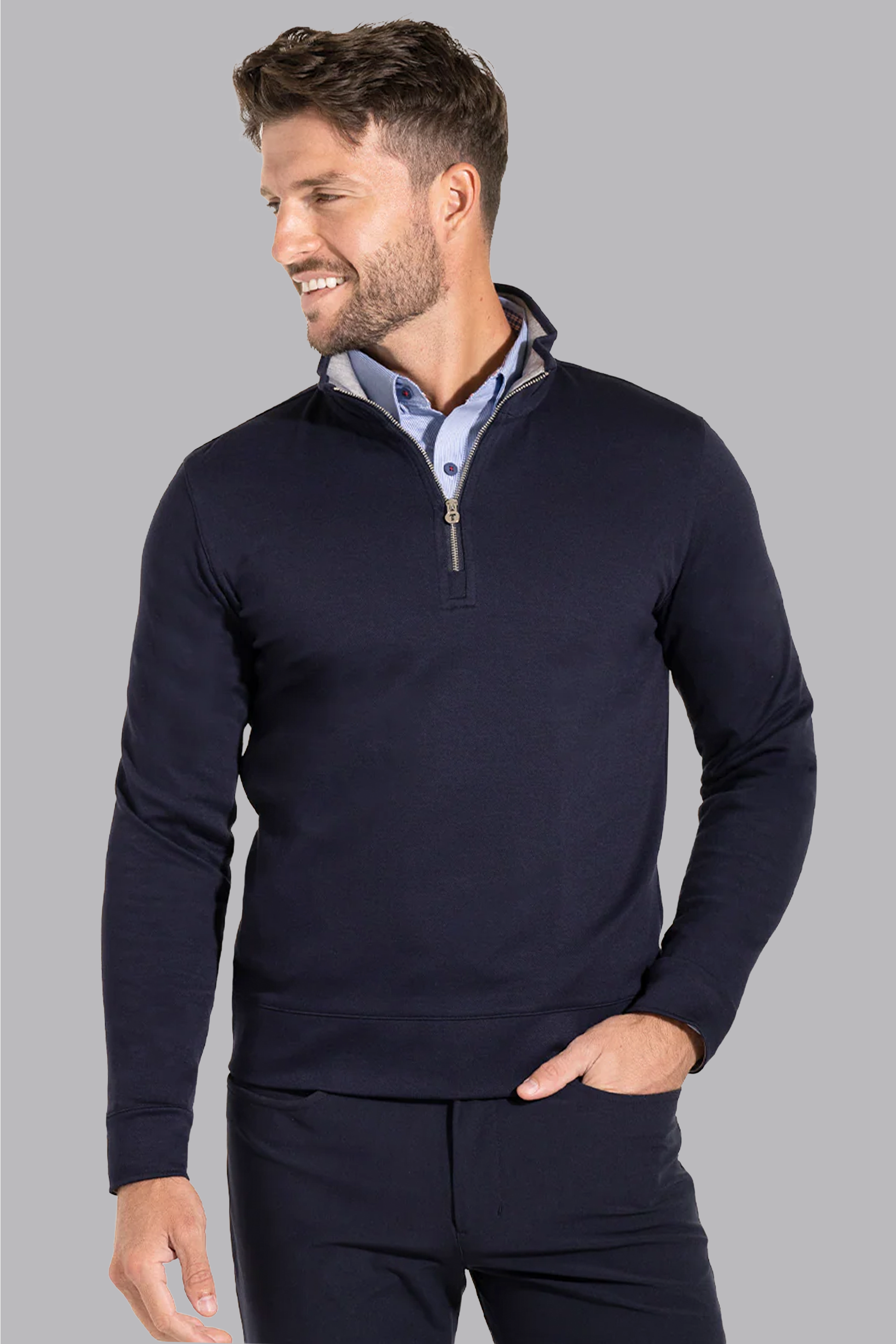 A man with short brown hair and a beard smiles, looking left, wearing the Twillory Performance Quarter Zip in Navy over a light blue collared shirt. One hand is in his pocket. The background is plain light gray—smart business casual style.