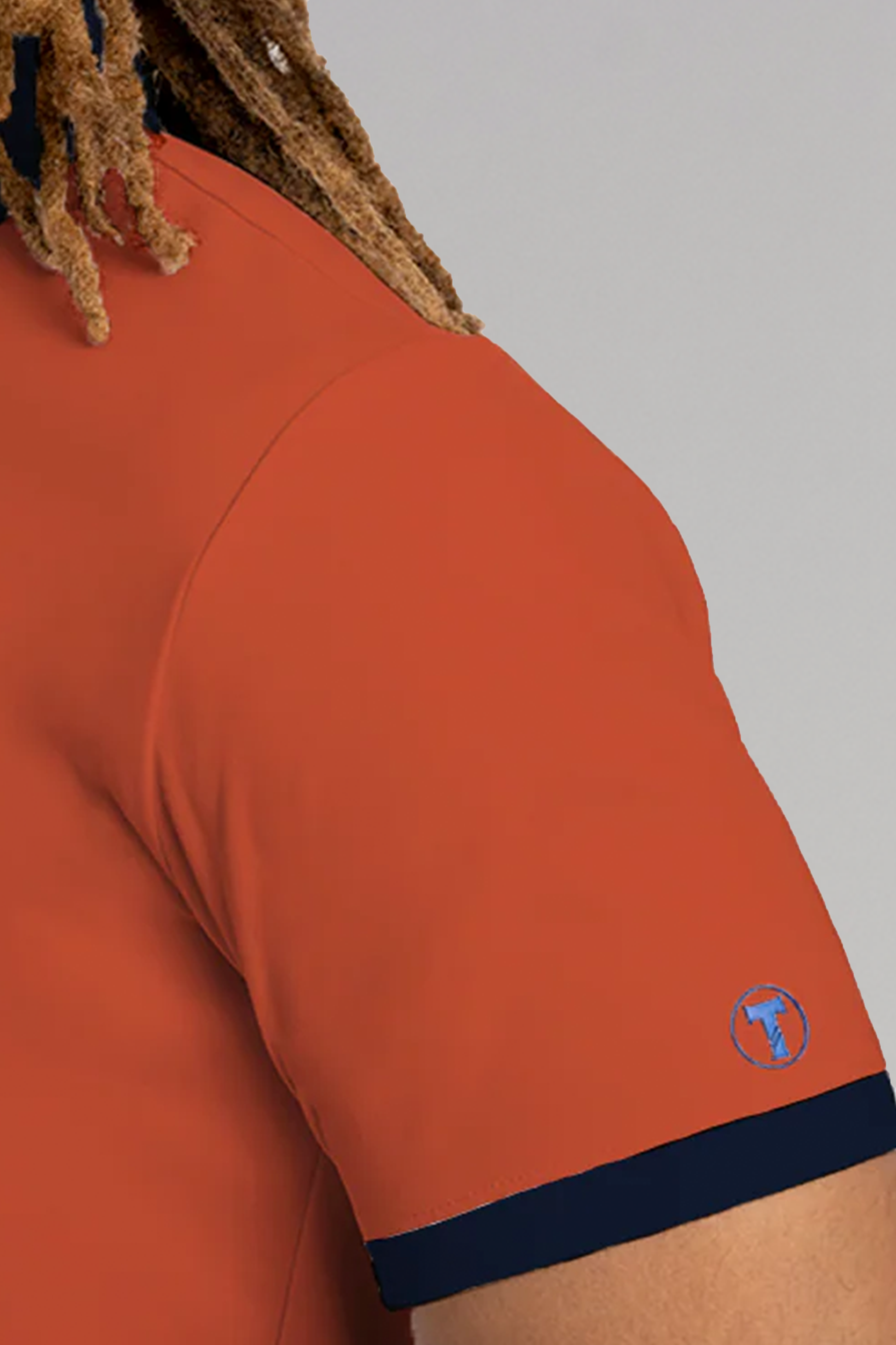 A close-up of a person’s upper arm in a Twillory Short Sleeve Polo Contrast in orange and navy, featuring black cuff trim and a small round blue logo with a white bull’s head on the sleeve. Brown, curly hair appears at the top left.