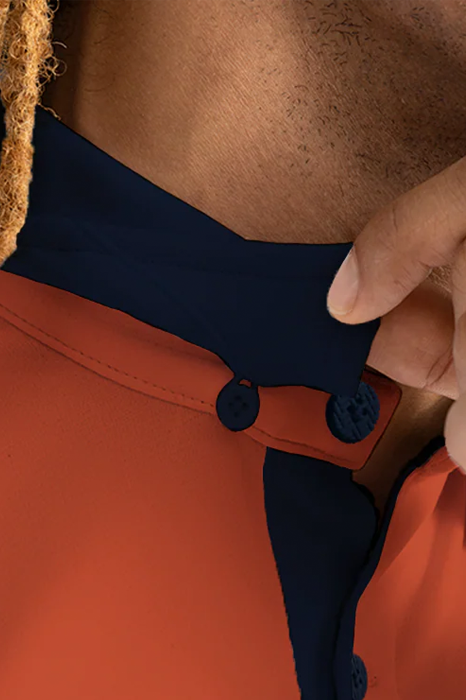 A close-up shows a person buttoning the Twillory Short Sleeve Polo Contrast // Orange Navy, featuring a burnt orange body, navy collar, dark blue trim, and buttons. Their brown skin and blond-brown dreadlock are visible. Cooling Tech ensures all-day comfort.
