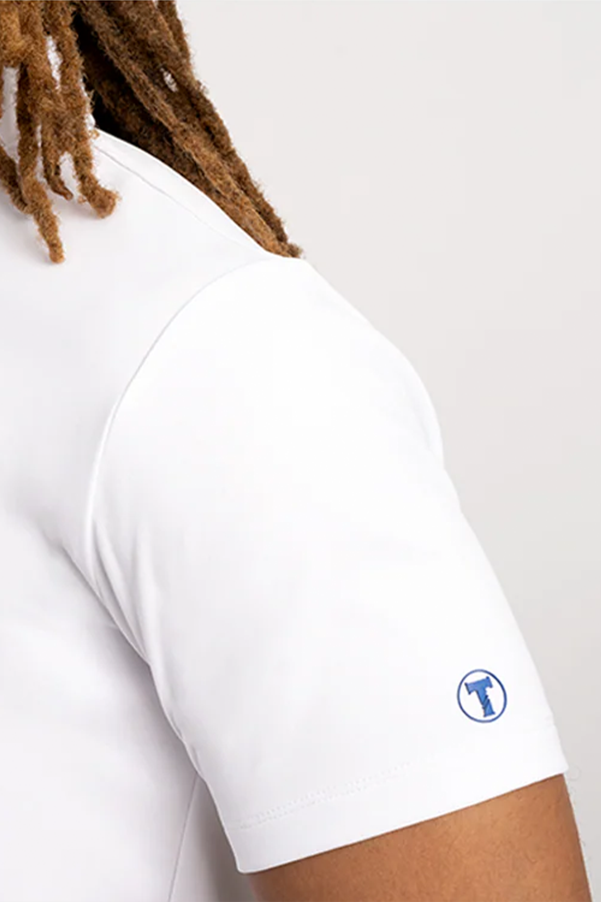 Close-up of a person in the Twillory Short Sleeve Polo Contrast // White Blue, featuring a tailored fit. The left sleeve shows a small blue circular logo with a t-shirt icon. Brown dreadlocks are partly visible; background is plain and light-colored.