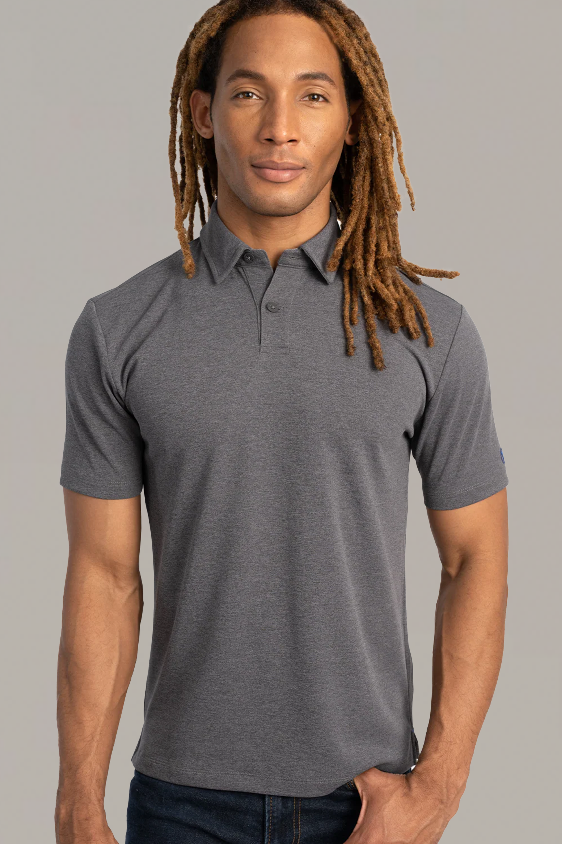 A person with long, light brown dreadlocks wears the Twillory Short Sleeve Polo Solid in Charcoal and blue jeans, standing against a plain light grey background and facing forward with a neutral expression.