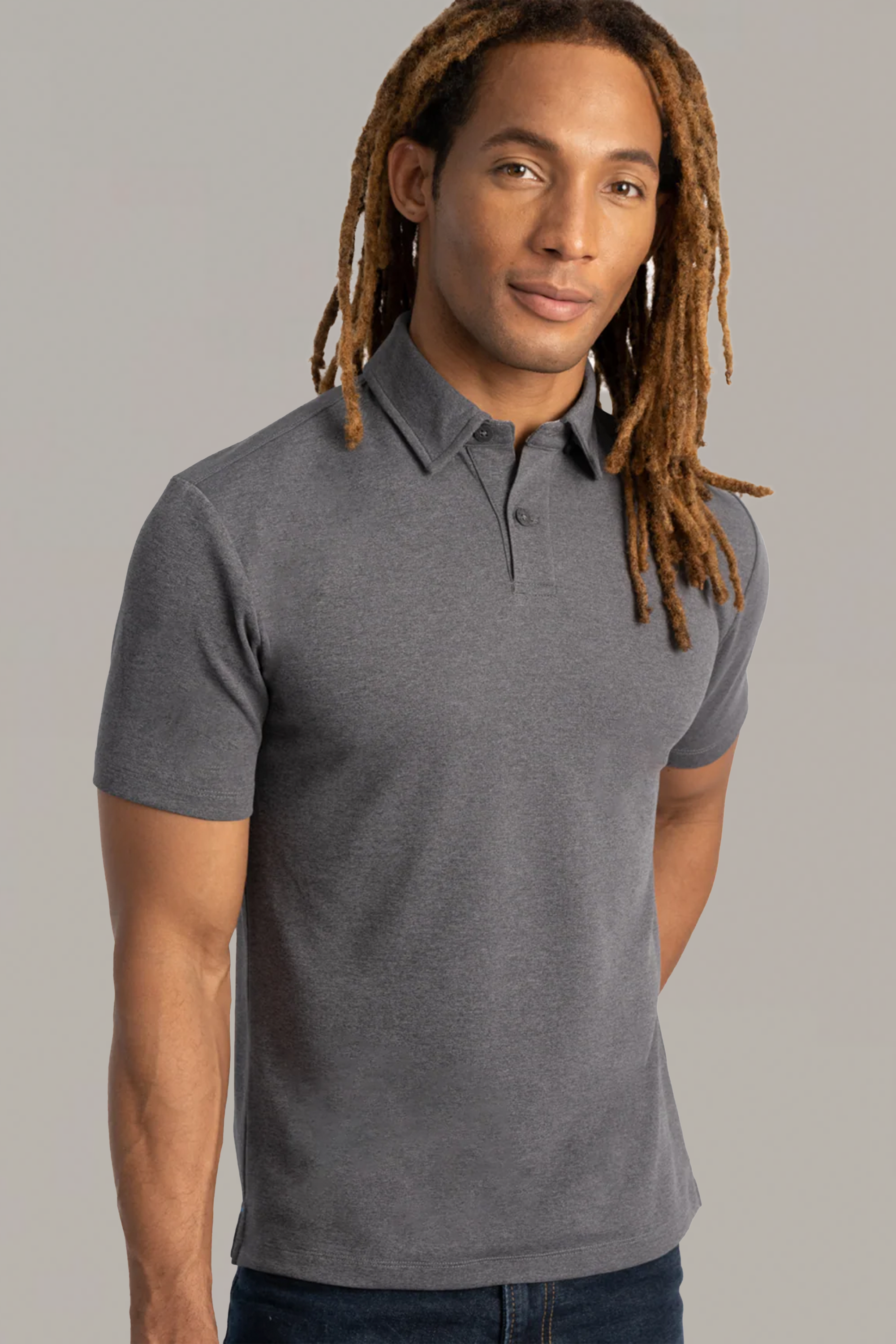 A person with long brown dreadlocks wears the Twillory Short Sleeve Polo Solid in Charcoal and dark jeans, standing against a plain light gray background and looking slightly to the side with a neutral expression.
