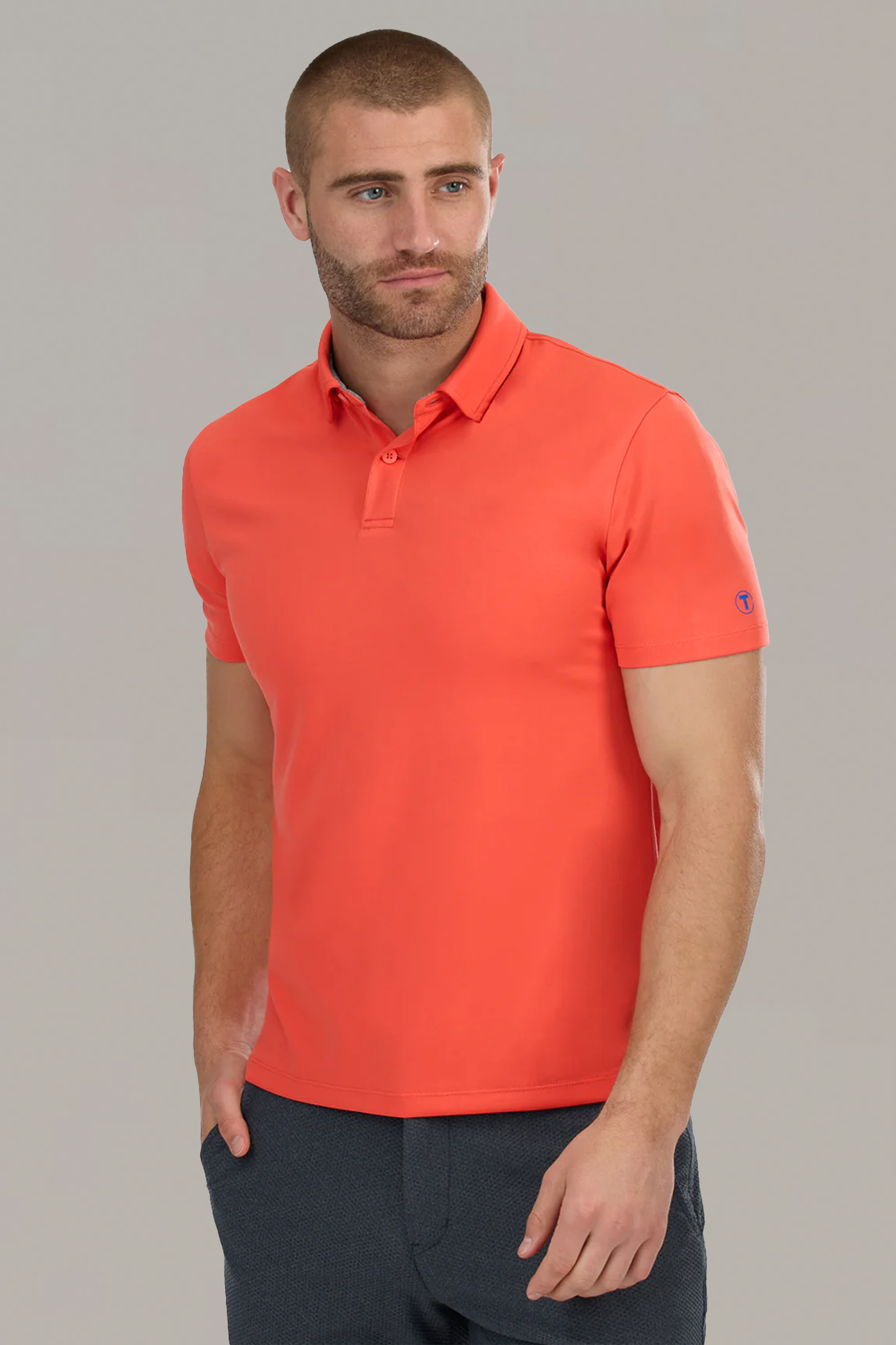 A man with a close-cropped beard and short hair wears the Twillory Short Sleeve Polo Solid in Salmon, featuring a tailored fit, collar, and short sleeves. With his left hand in his pocket, he stands against a plain light gray background, looking slightly right.