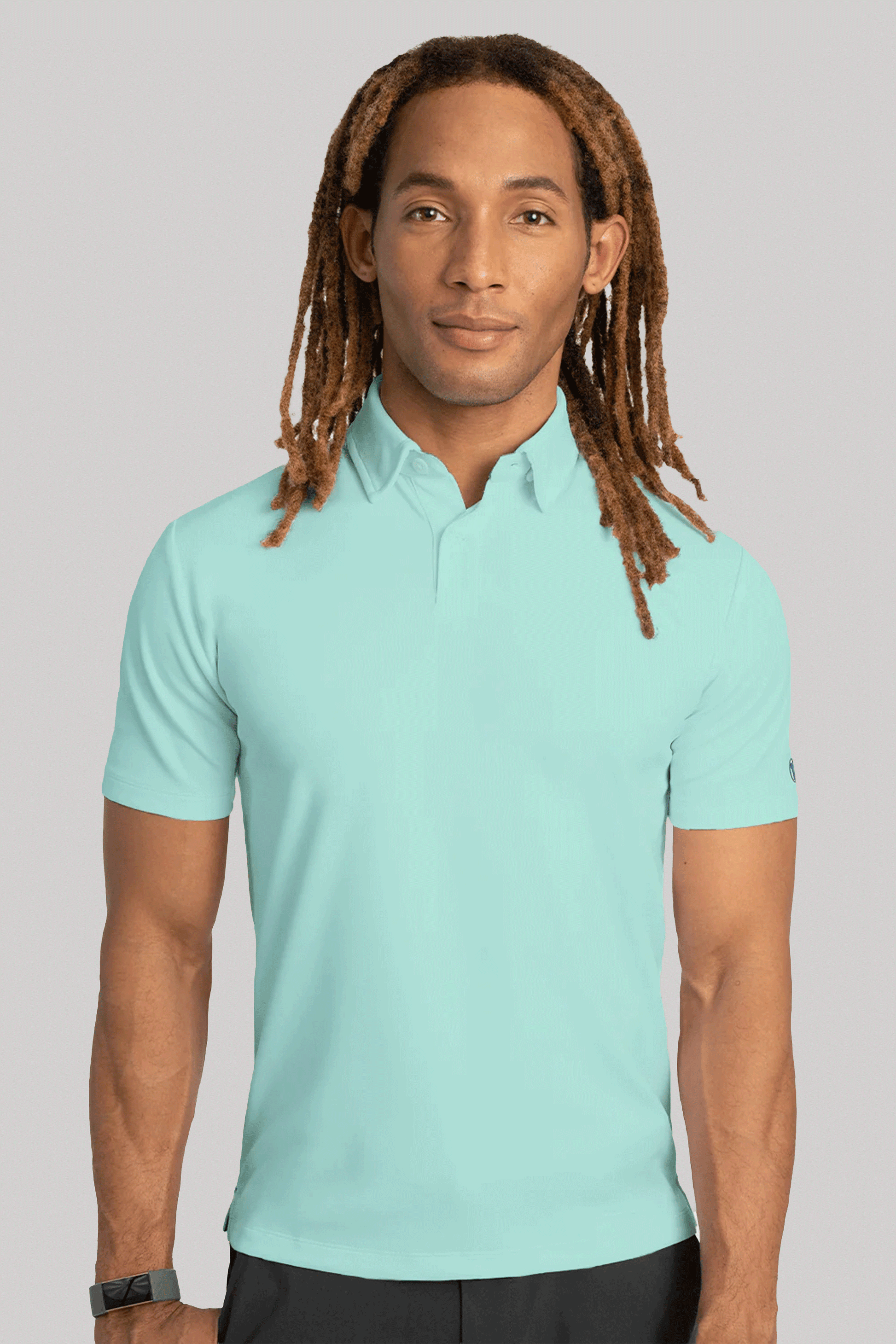 A man with long, light brown dreadlocks faces the camera wearing a Twillory Short Sleeve Polo Solid in Turquoise and black pants. He has a neutral expression, a black wristwatch on his left wrist, and stands against a plain light gray background.