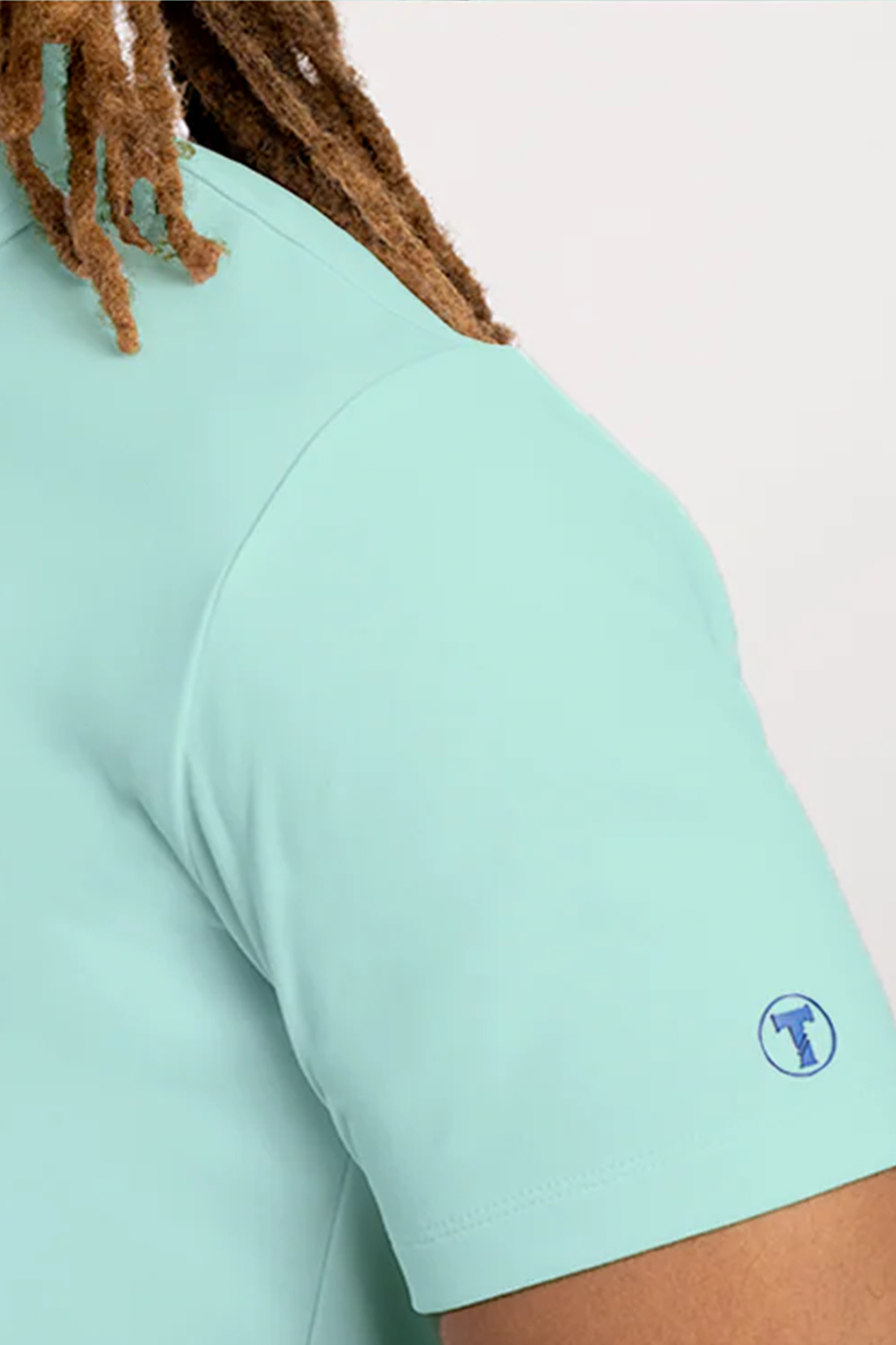Close-up of a person wearing the Twillory Short Sleeve Polo Solid in turquoise. A small blue circular logo with a T-shirt icon appears near the sleeve hem, and part of their brown, twisted hair is visible against a plain light background.
