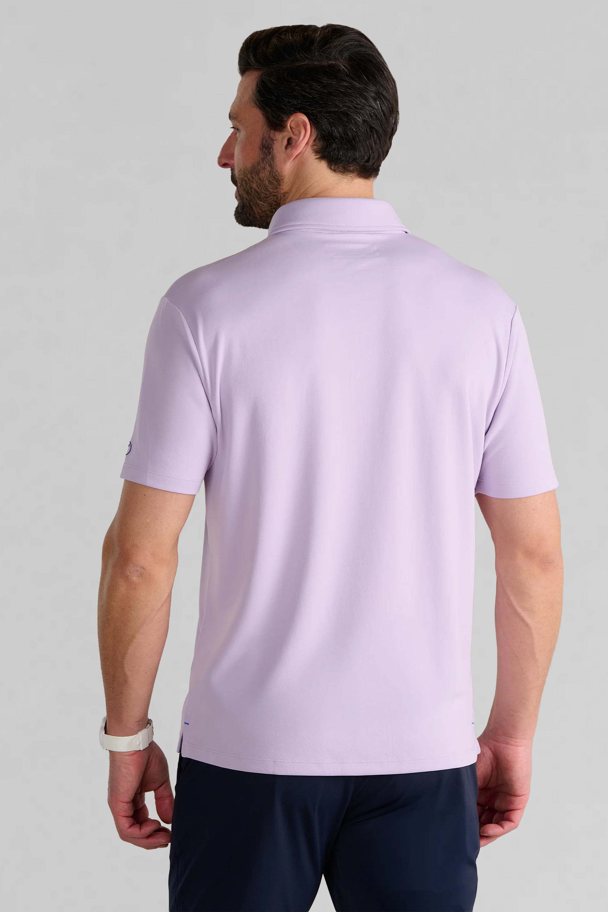 A man with short dark hair and a beard stands with his back to the camera, wearing the Twillory Short Sleeve Polo Solid in Lilac and dark navy blue pants. He has a white watch on his left wrist, set against a plain light gray background.