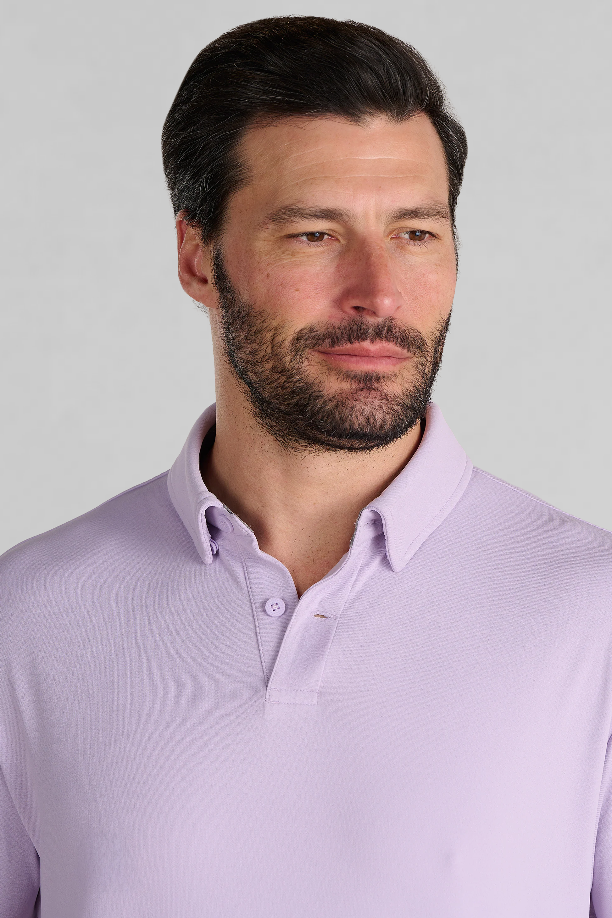 A man with short dark hair and a beard wears the Twillory Short Sleeve Polo Solid in Lilac. He looks slightly left with a neutral expression, standing against a plain light gray background that highlights his moisture-wicking shirt.