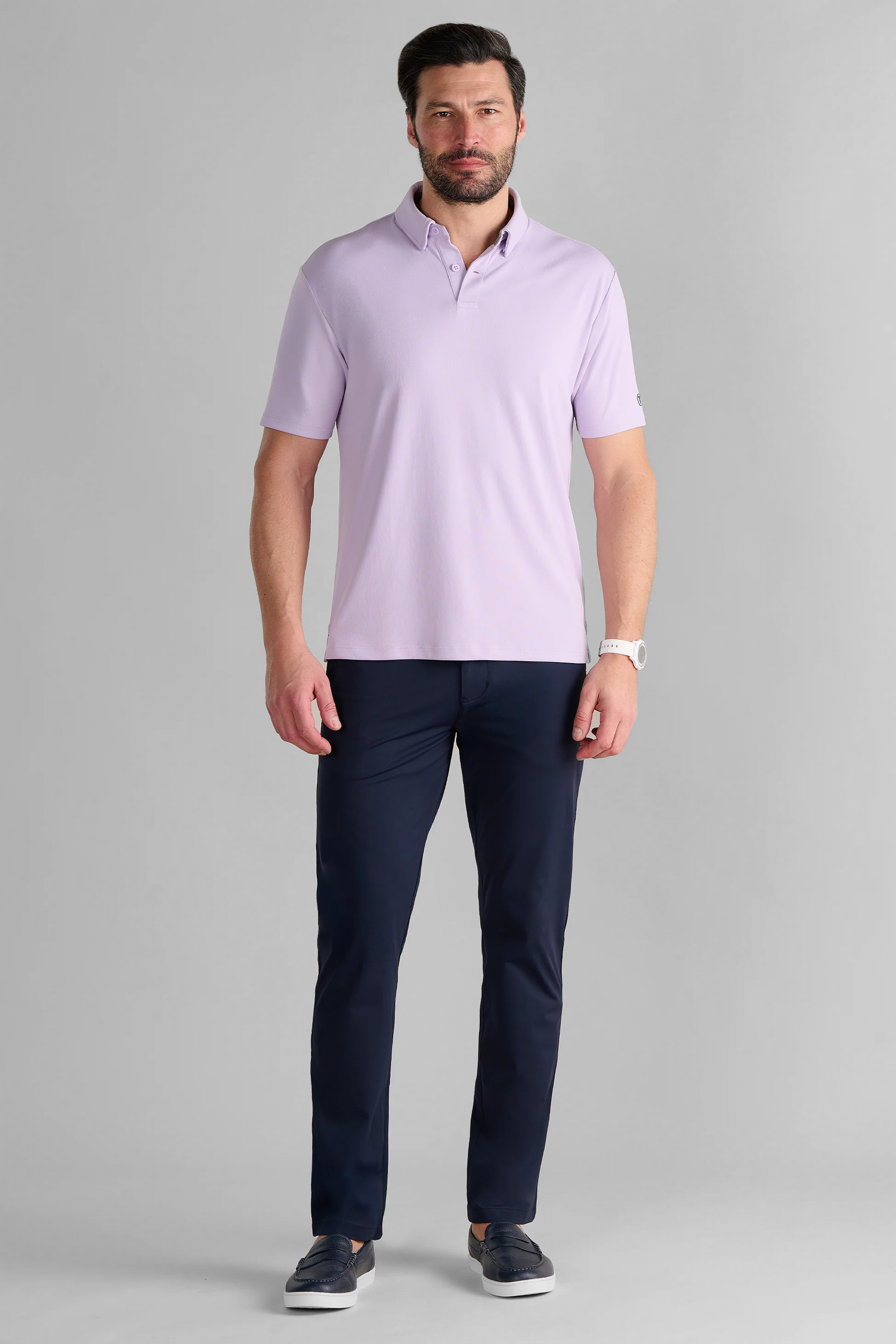 A man with short dark hair and a beard stands facing forward, wearing the Twillory Short Sleeve Polo Solid in Lilac, paired with dark navy pants, navy slip-on shoes, and a white watch. He poses against a plain gray background with arms relaxed at his sides.