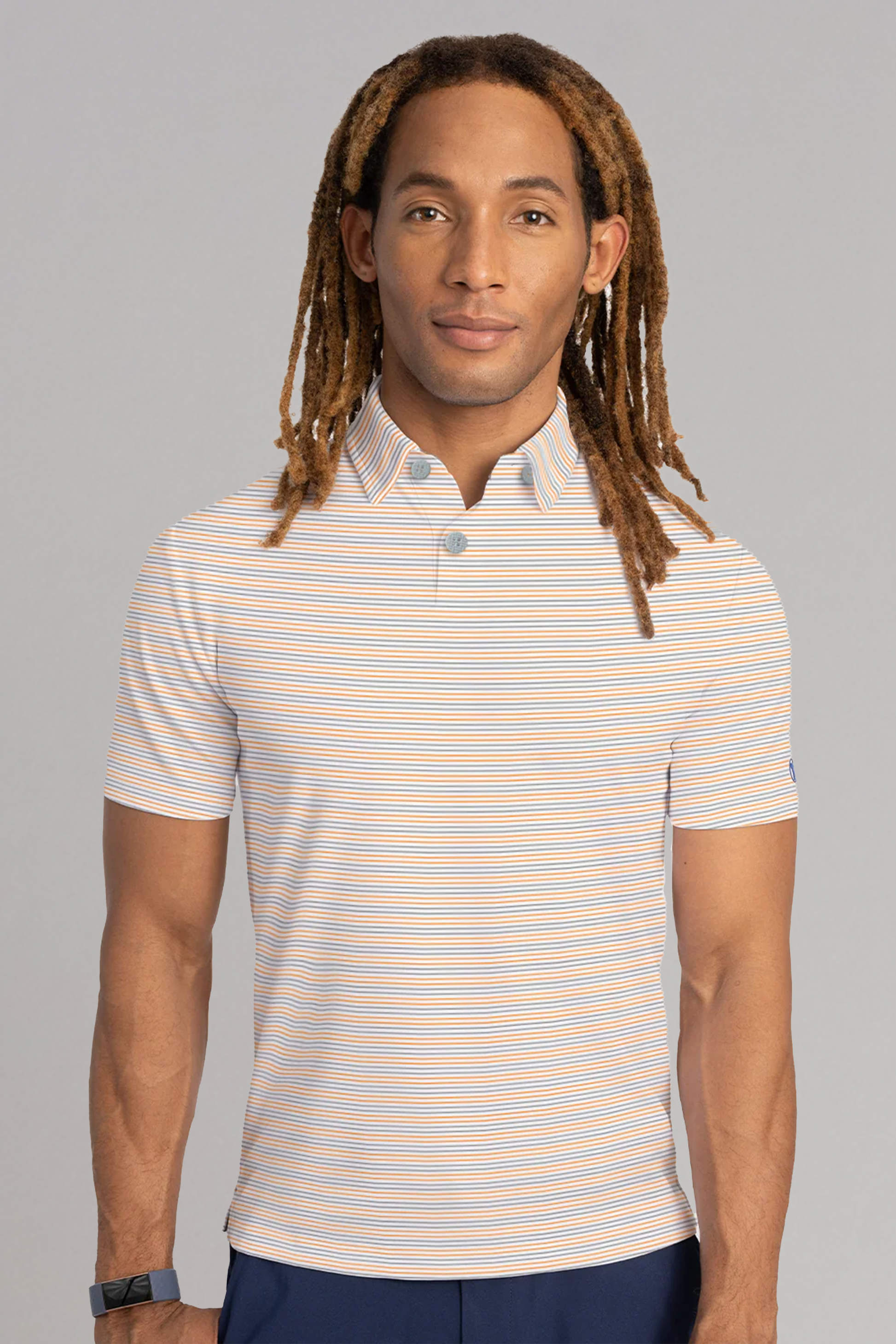 A man with long, light brown dreadlocks faces forward, wearing the Twillory Short Sleeve Polo Stripe // Grey Orange and dark pants. He has a smart watch on his left wrist. The background is plain gray.