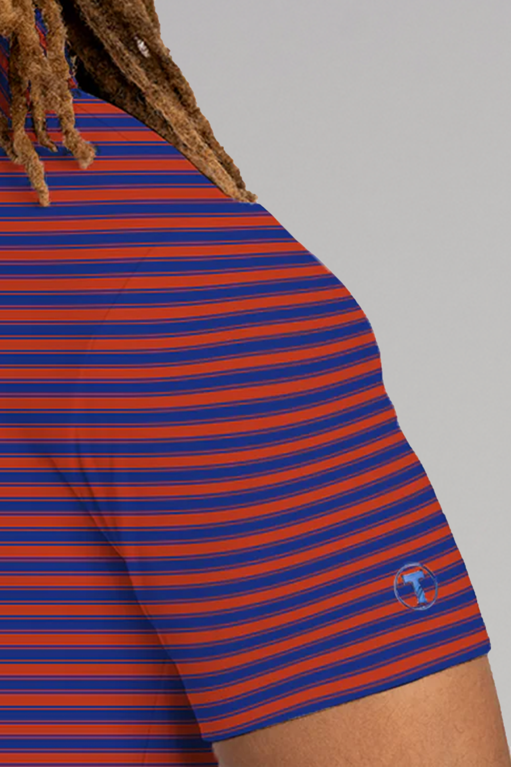 A close-up shows a person's shoulder in the Twillory Short Sleeve Polo Stripe // Red Blue, featuring blue and red stripes, twisted brown hair, and a small embroidered sleeve logo against a light gray background.