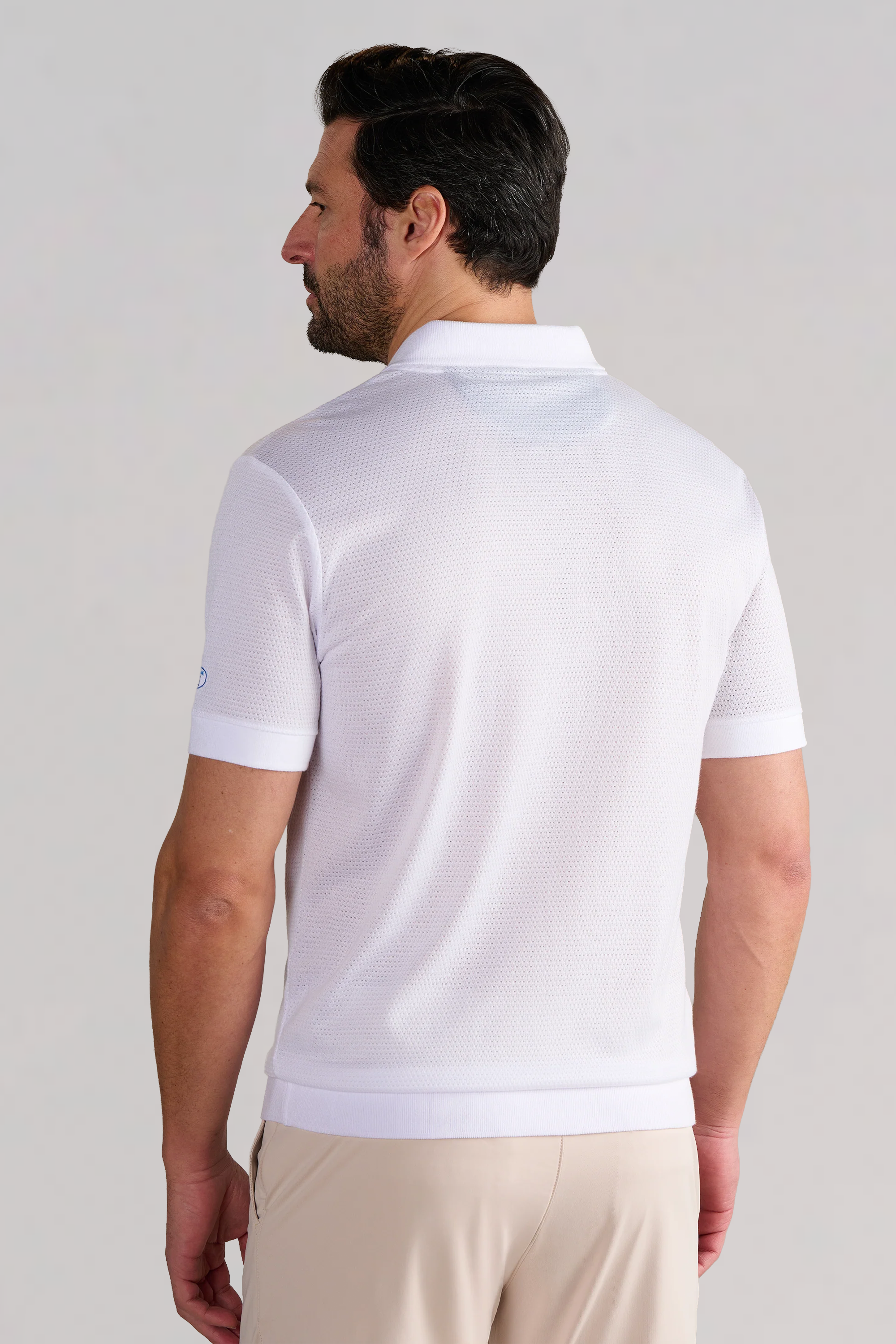 A man with short dark hair and a trimmed beard is seen from behind wearing Twillory’s Short Sleeve Sweater Polo Solid in White, featuring textured fabric and ribbed cuffs, paired with beige pants against a plain light gray background.