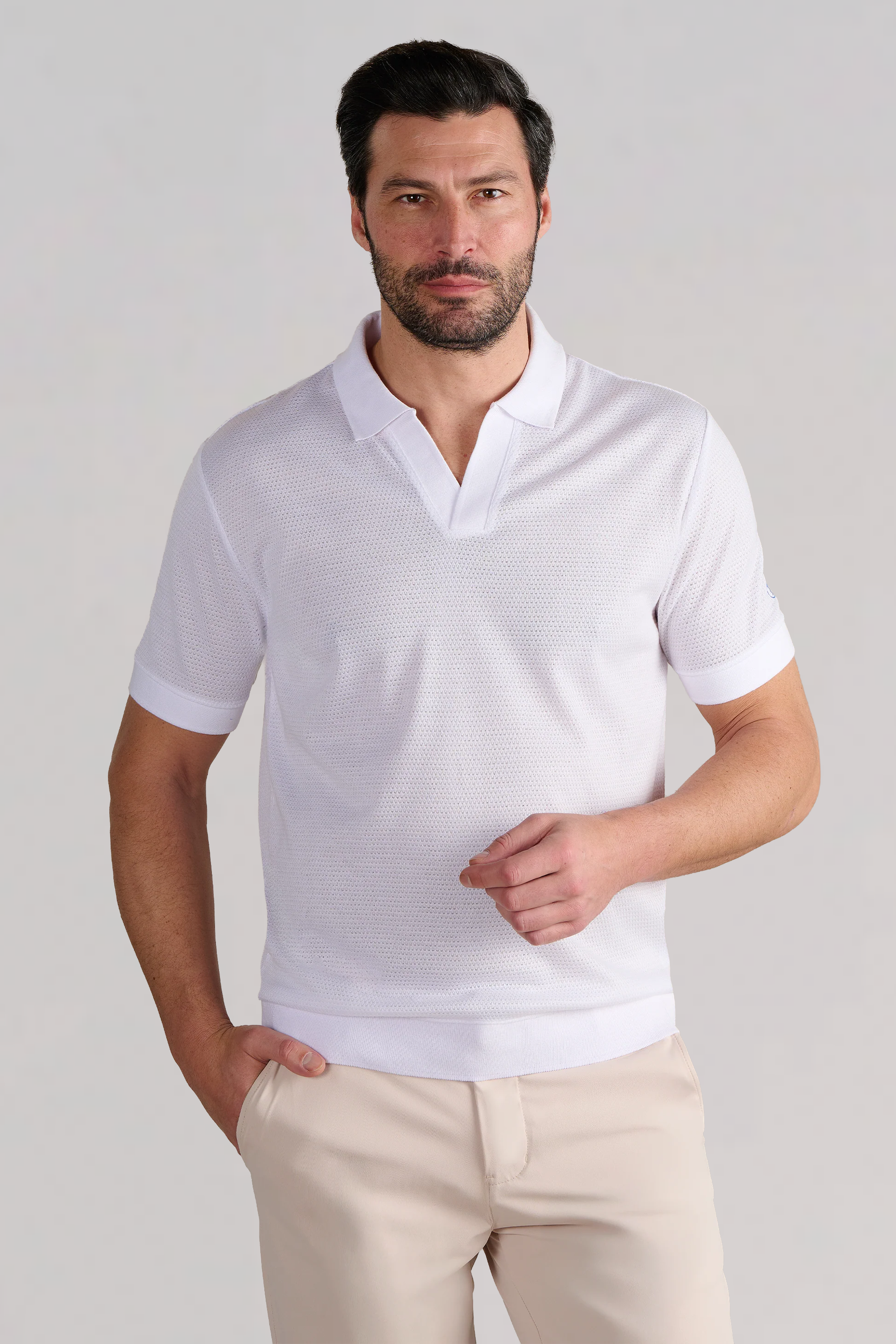 A man with short dark hair and a trimmed beard wears the Twillory Short Sleeve Sweater Polo Solid in white and beige pants. He stands against a light gray background, one hand in his pocket, looking straight ahead.