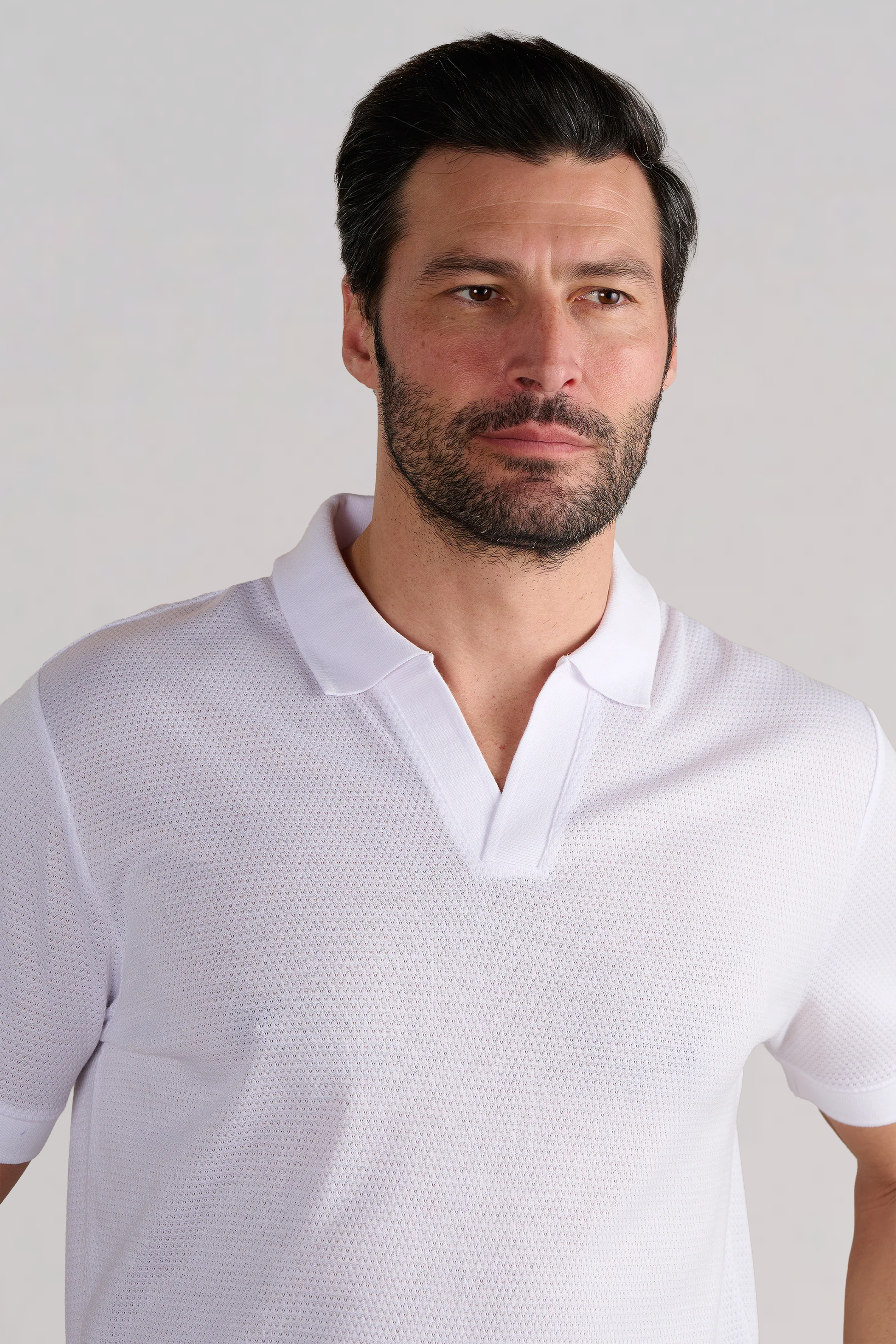 A man with short dark hair and a beard wears the Twillory Short Sleeve Sweater Polo Solid in White, a wrinkle-free performance knit with a split collar. He looks to the side with a neutral expression against a plain light background.