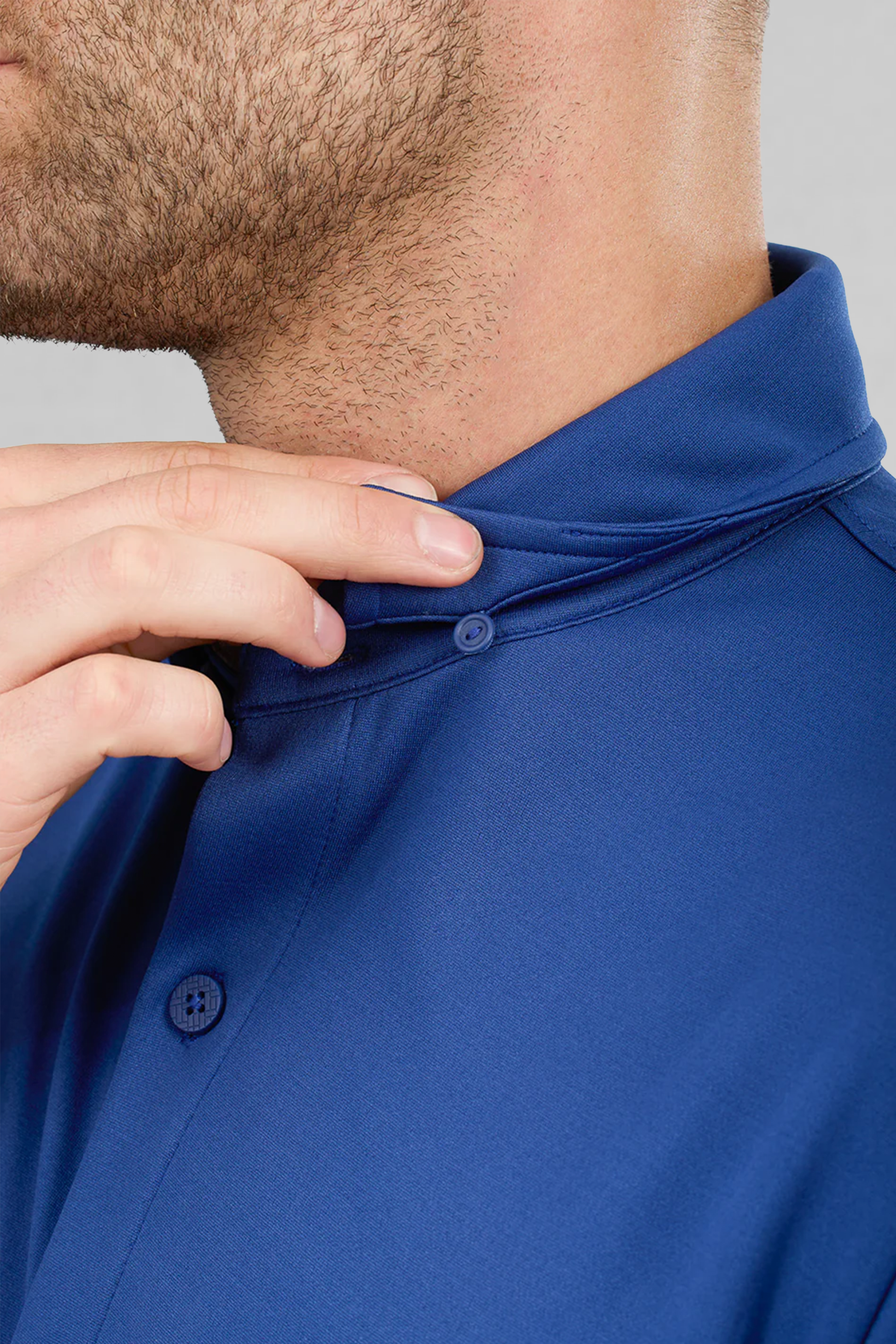 A light-skinned man with a trimmed beard uses his left hand to fasten a hidden button under the collar of a Twillory Short Sleeve Button Down Polo Solid in Twilight, made from smooth, moisture-wicking, wrinkle-free fabric. The background is plain light gray.