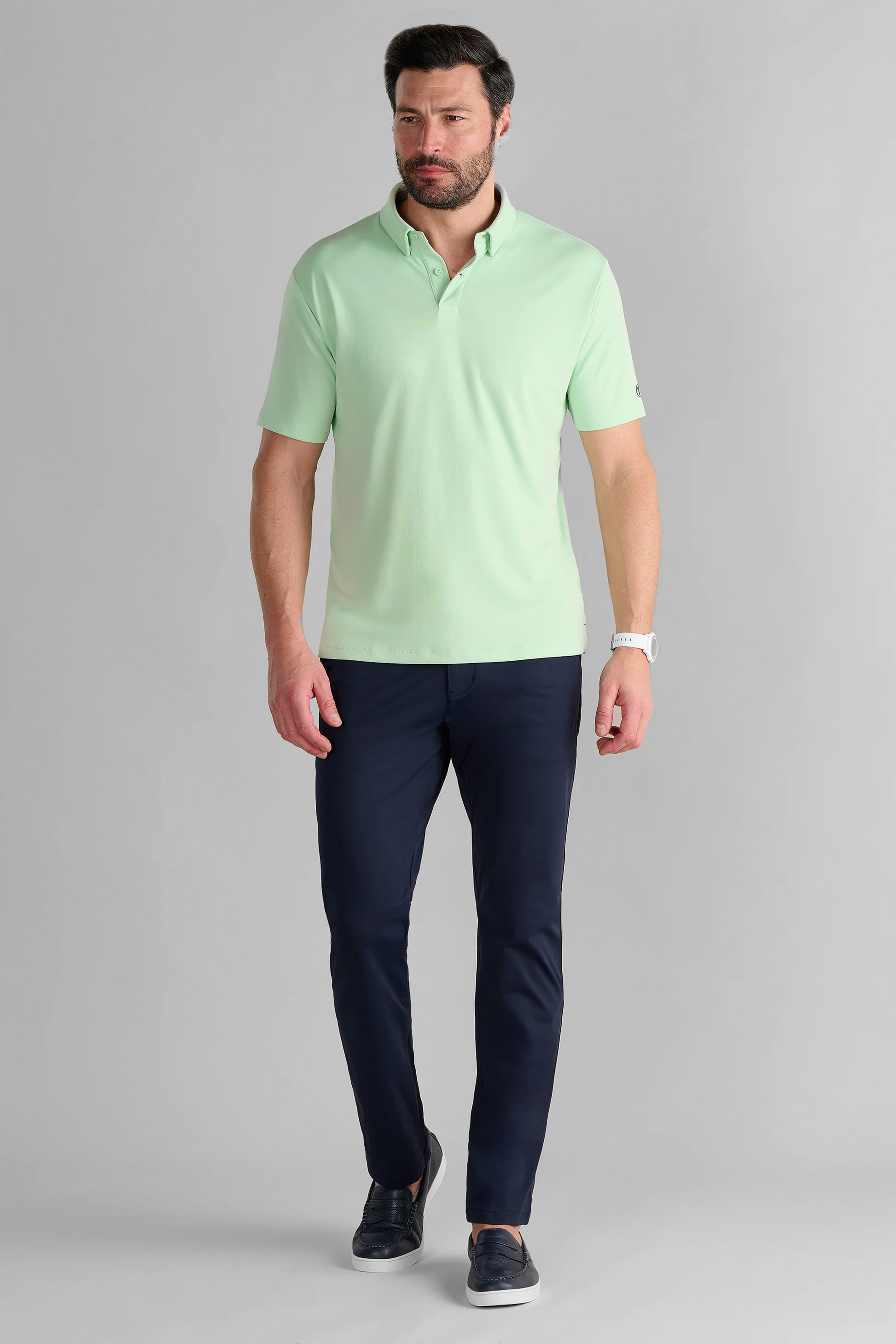 A man with short dark hair and a beard wears the Twillory Short Sleeve Polo Solid in Pastel Green, navy blue pants, and dark slip-on shoes. He stands mid-step against a plain gray background as if walking.
