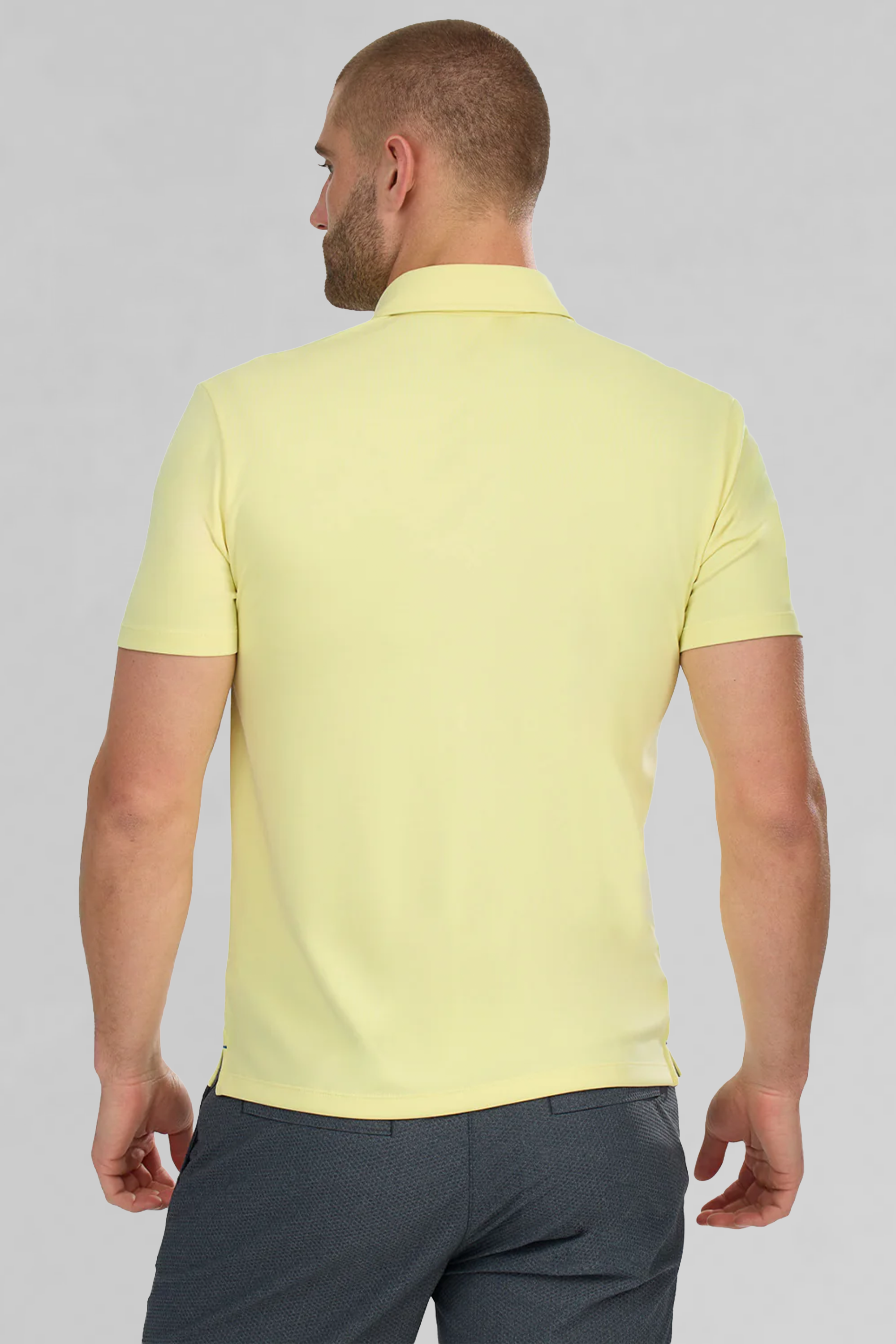 A man with short hair, seen from behind, wears the Twillory Short Sleeve Polo Solid in yellow and dark gray pants. He stands against a plain light gray background with his arms relaxed at his sides.