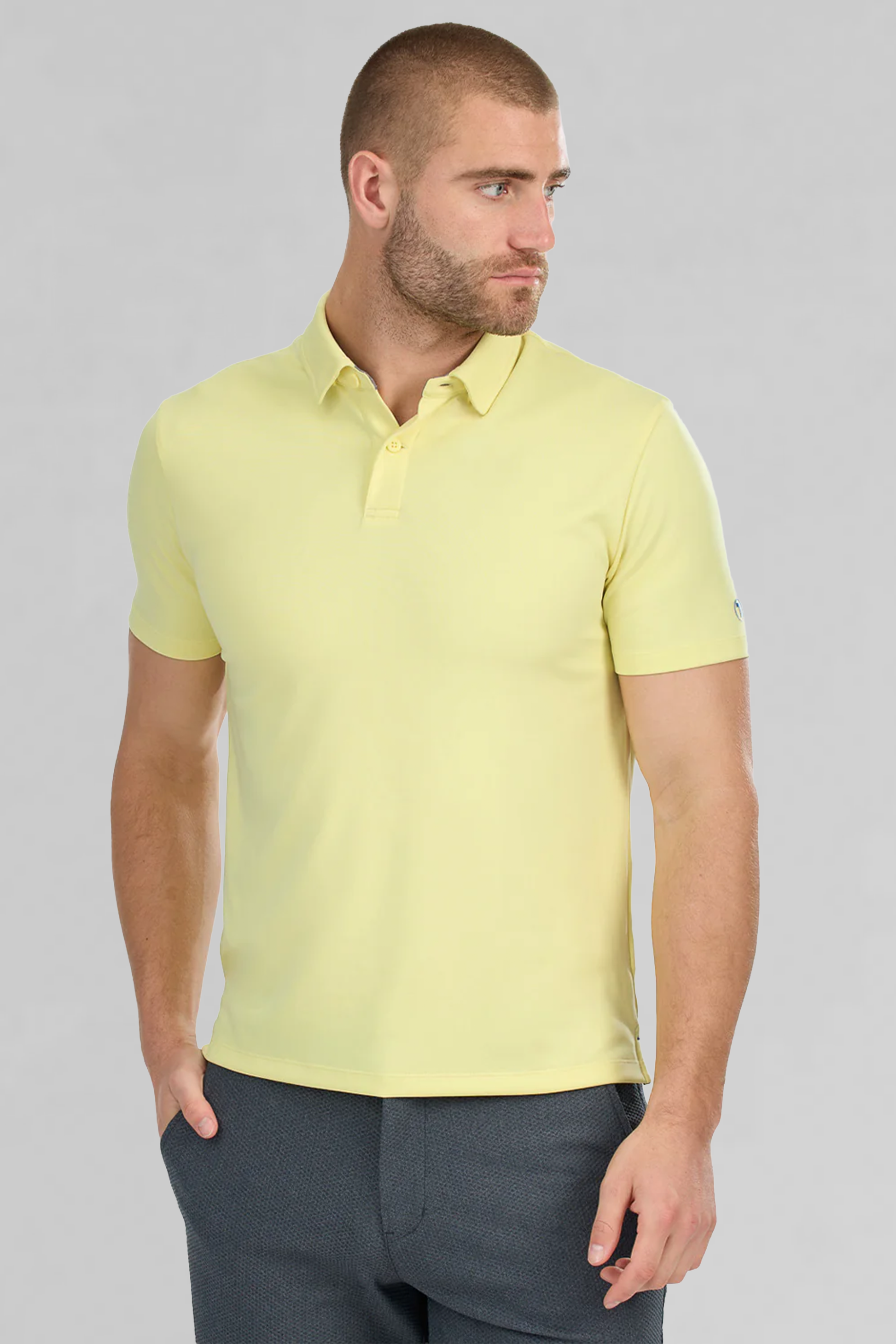 A man with short hair and a trimmed beard wears the Twillory Short Sleeve Polo Solid in yellow and dark trousers, standing with one hand in his pocket and looking left against a plain light gray background.