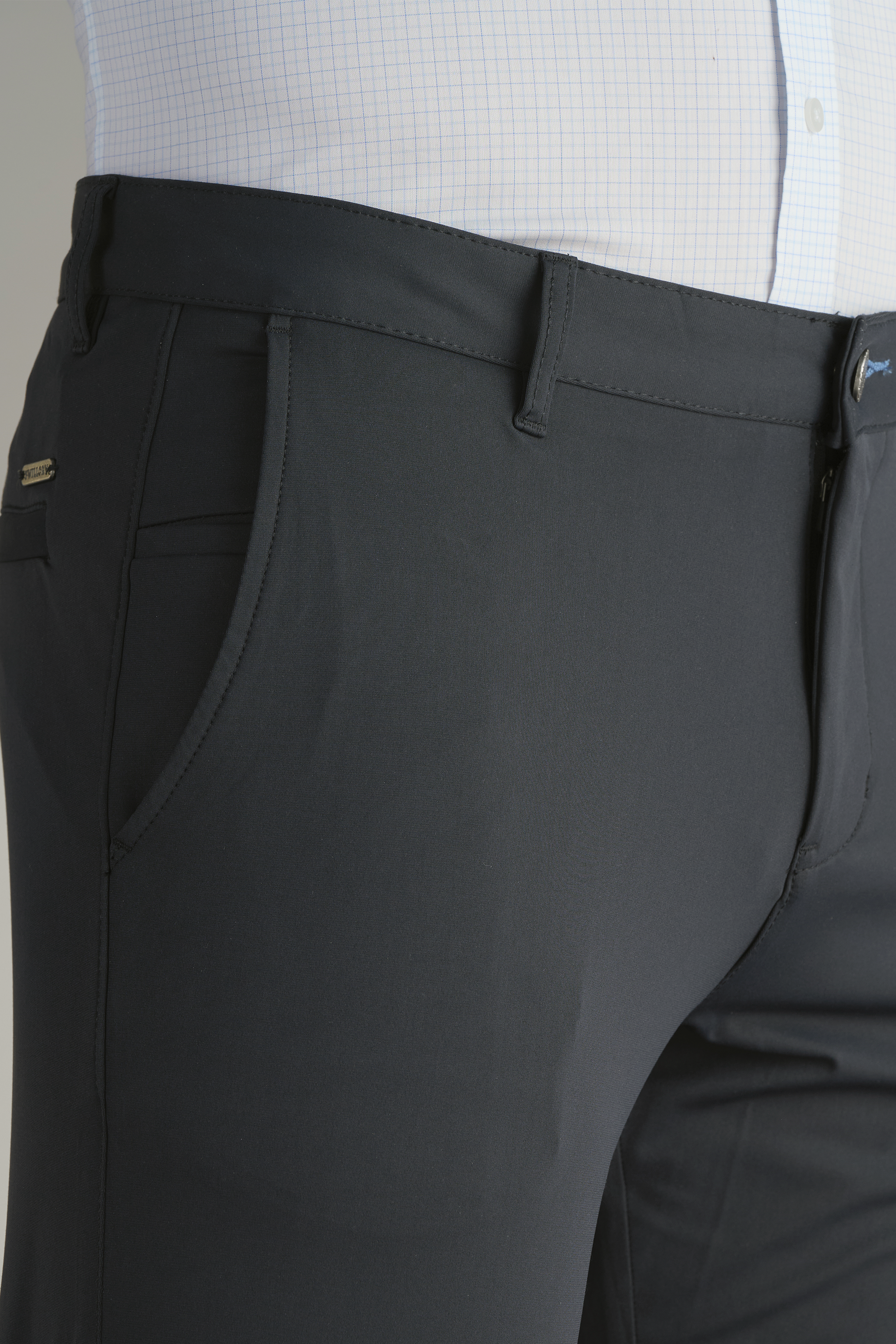 A close-up shows a person in Twillory Performance Pants // SLATE GREY with front pockets and belt loops, paired with a tucked-in light blue checkered shirt. Only the torso and upper legs are visible against a plain background.