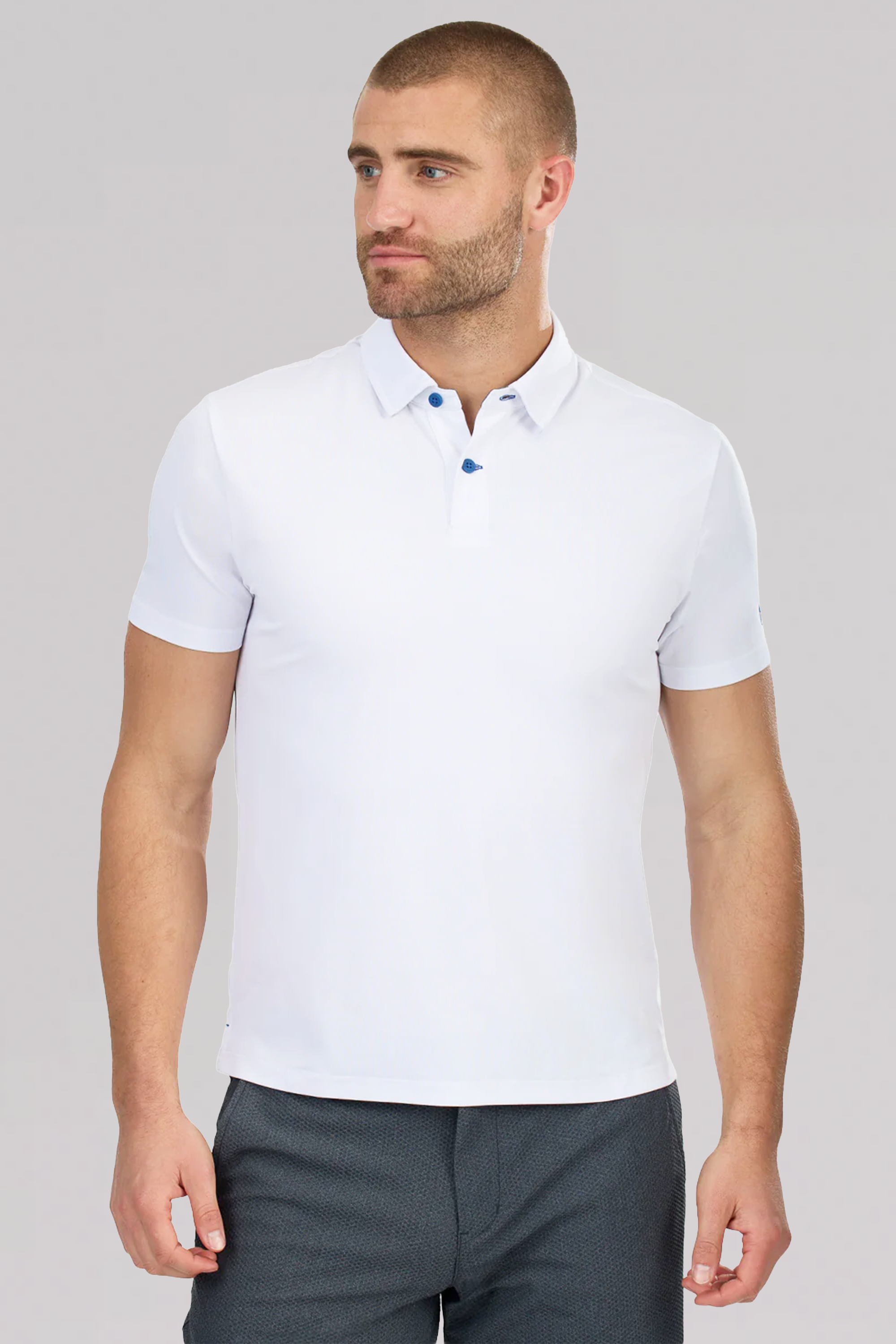 A man with short brown hair and a beard models the Twillory Slim Fit Pique Polo Contrast in White Blue, featuring blue buttons, paired with dark gray pants. He stands against a plain light gray background, looking slightly to the side.