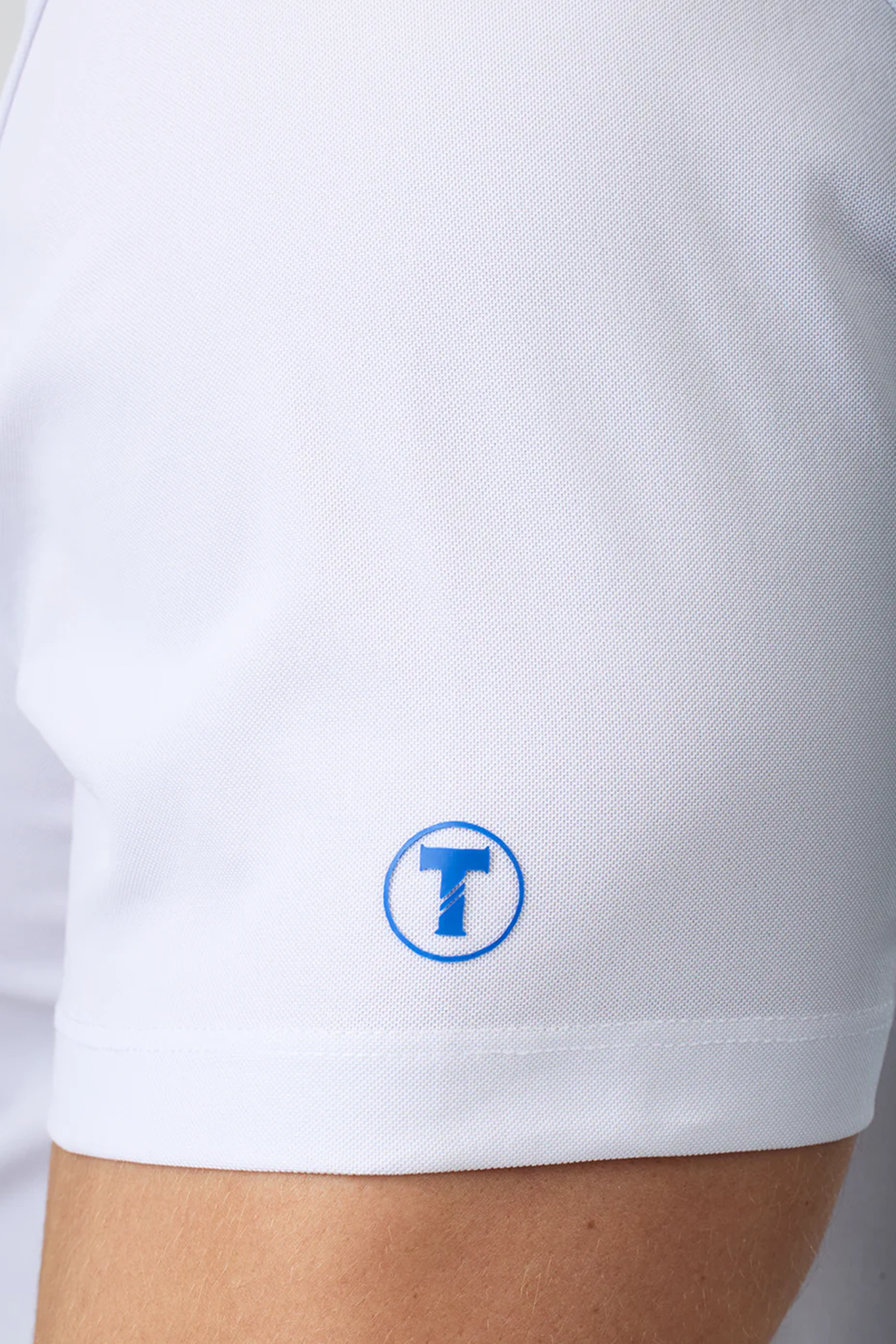 Close-up of a person wearing the Twillory Slim Fit Pique Polo Contrast in White Blue. A small blue and white circular logo with a bold "T" appears near the sleeve hem. The white, moisture-wicking polo stands out against the blurred background.