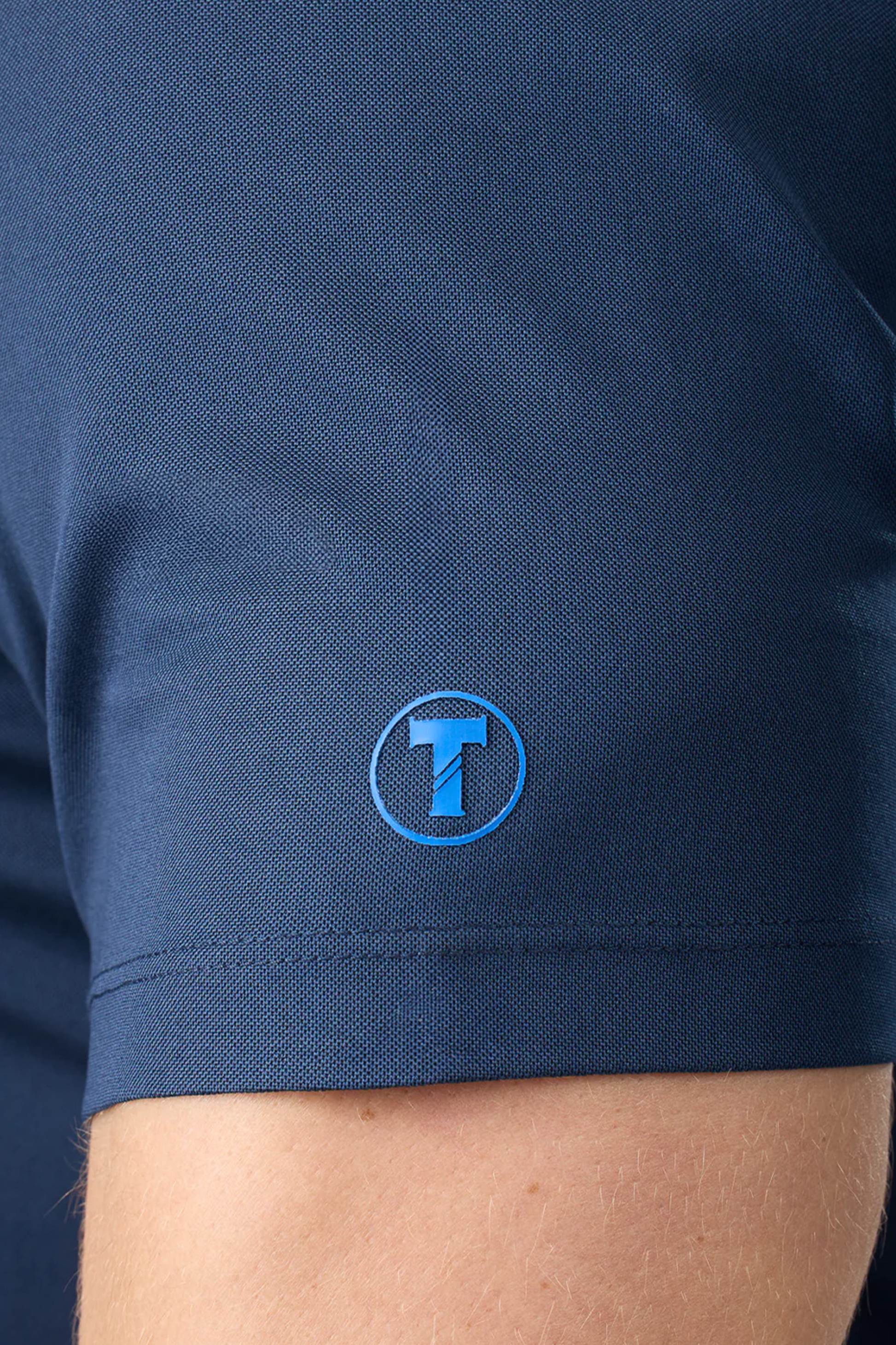 Close-up of a light-skinned person’s upper arm wearing Twillory’s Slim Fit Pique Polo Solid in Navy, featuring a small blue circular “T” logo near the sleeve edge.