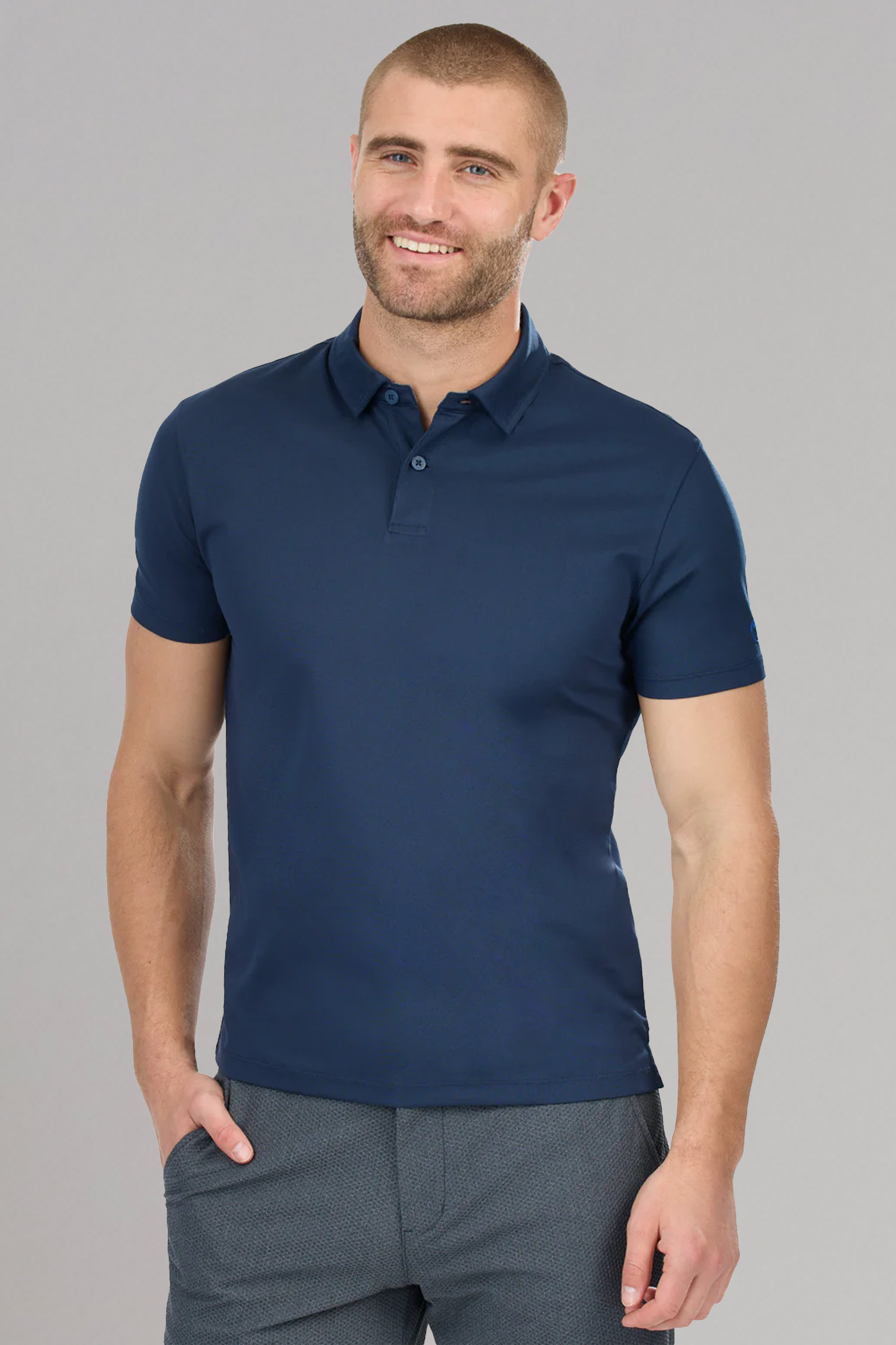 A man with short hair and a beard smiles in a Twillory Slim Fit Pique Polo Solid in navy, paired with textured gray pants, standing against a plain light gray background with one hand in his pocket.