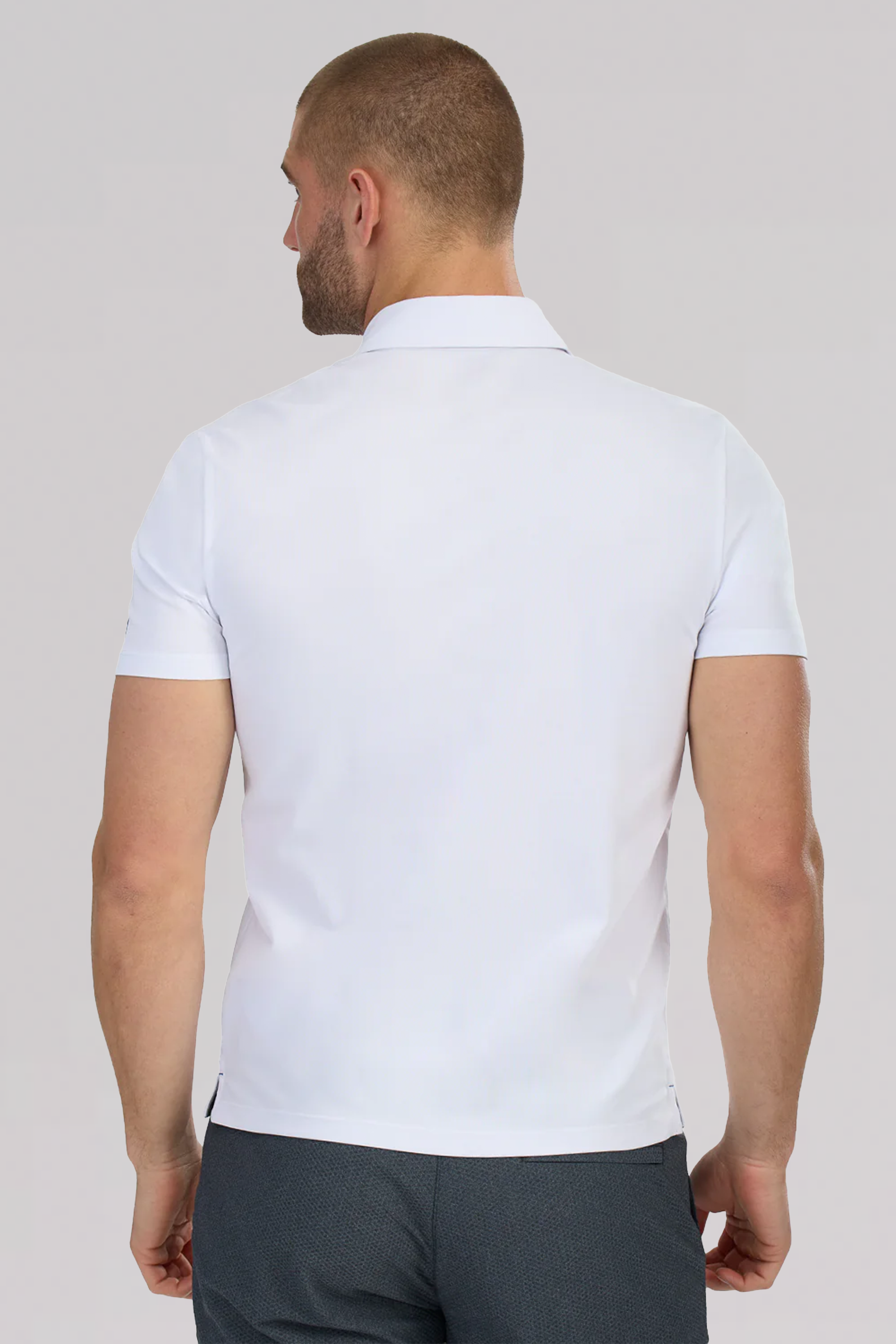 A man with closely cropped hair is shown from behind, wearing a white Twillory Slim Fit Pique Polo Solid and dark pants. The polo’s classic collar and snug fit accentuate his athletic build against a plain light gray background.
