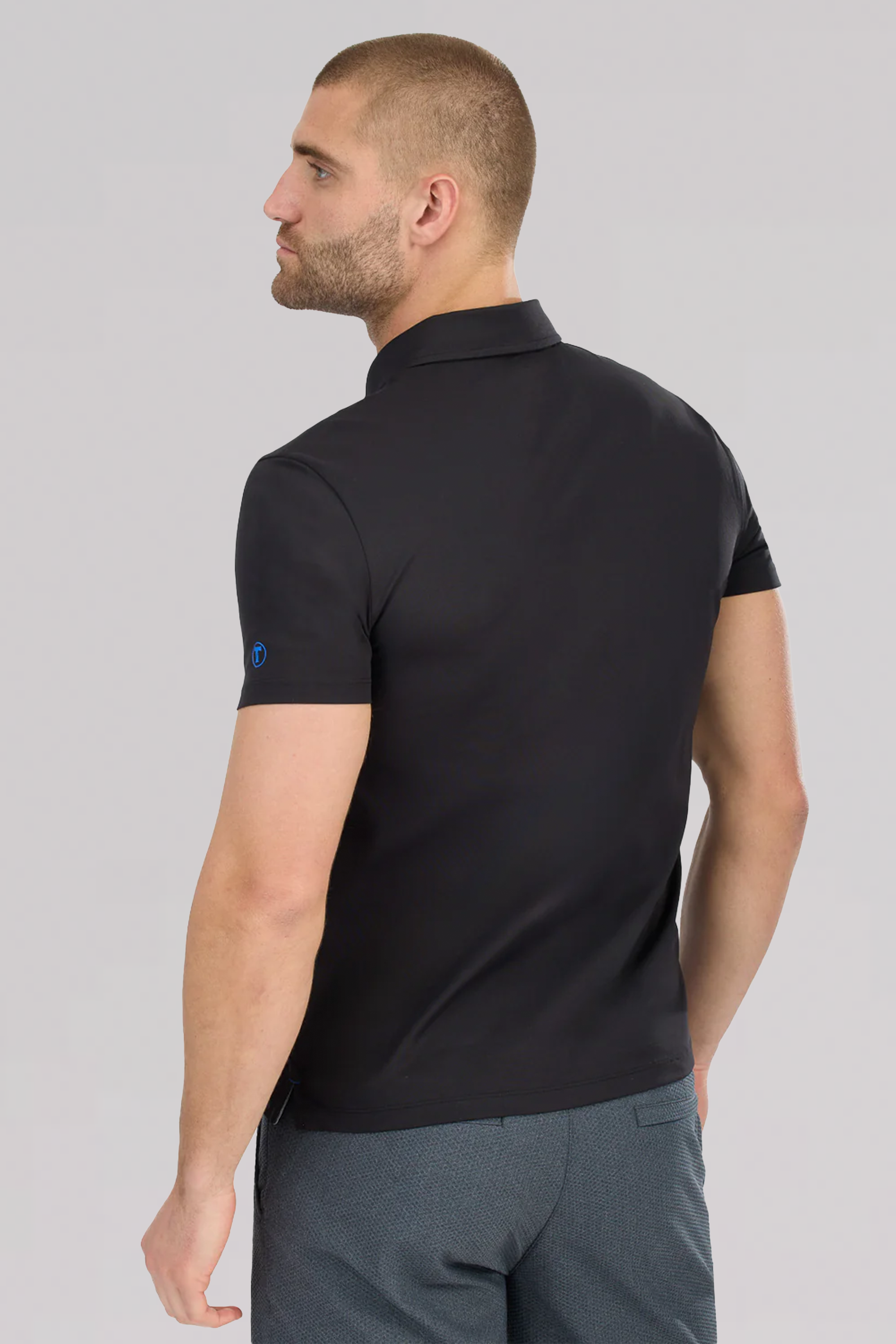 A man with short hair and a beard is seen from the back in a black Twillory Slim Fit Pique Polo Solid and gray textured pants. The polo has short sleeves, moisture-wicking fabric, and a subtle blue logo on one sleeve. The background is plain and light-colored.