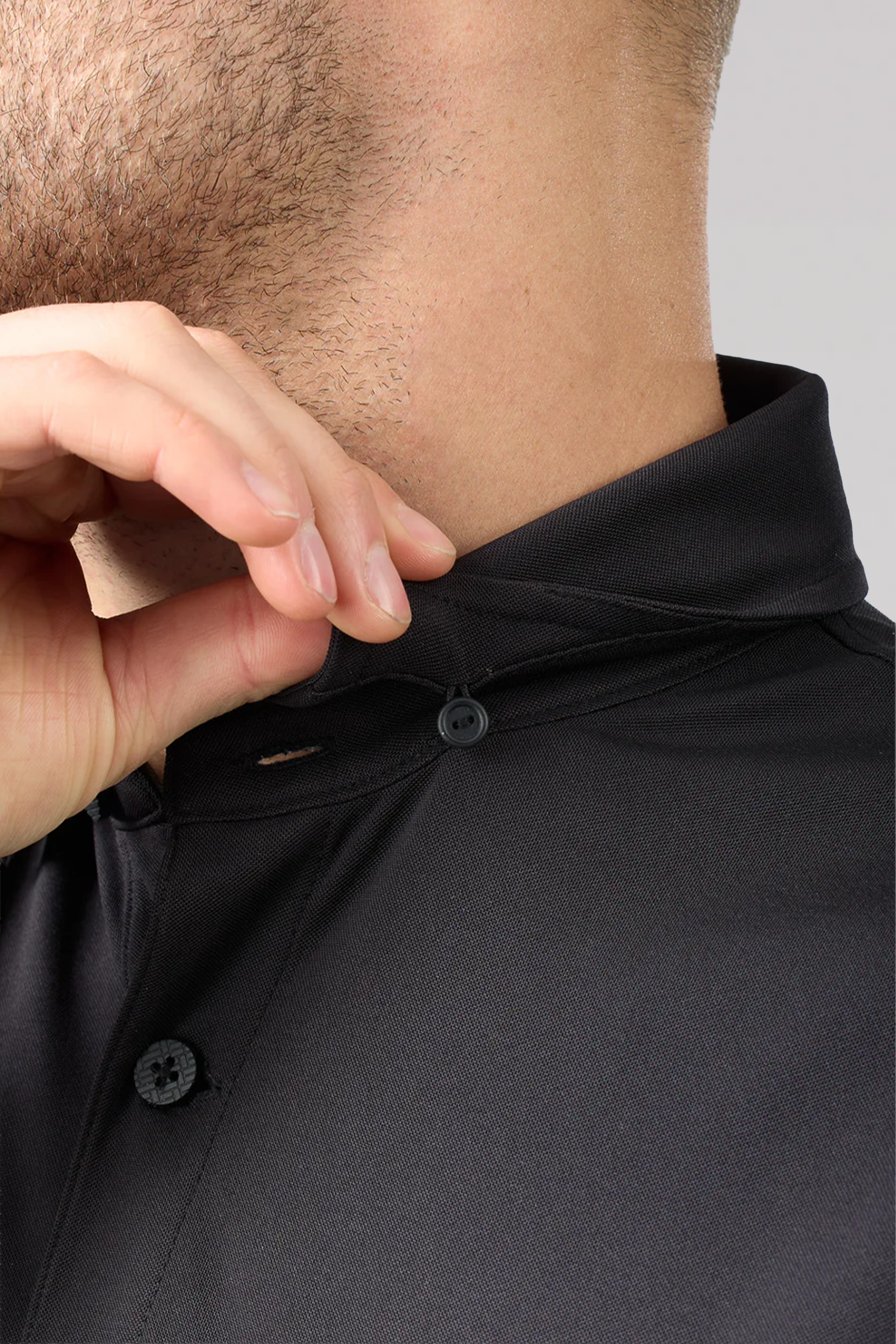A close-up shows a person buttoning the top of a Twillory Slim Fit Pique Polo Solid in Black, highlighting the smooth, slightly shiny, moisture-wicking fabric around the neck and chin.
