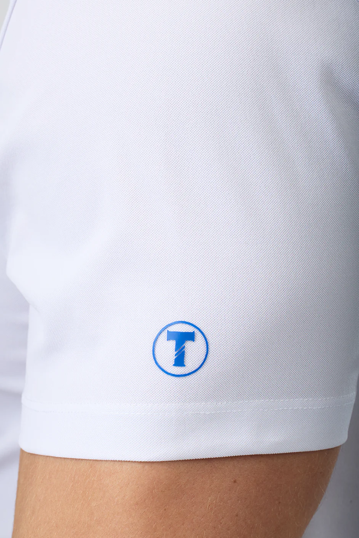 Close-up of a person wearing the Twillory Slim Fit Pique Polo Solid in white. A small circular blue and white logo with a stylized T appears on the sleeve, and the textured fabric adds both style and function. Part of the arm is visible.