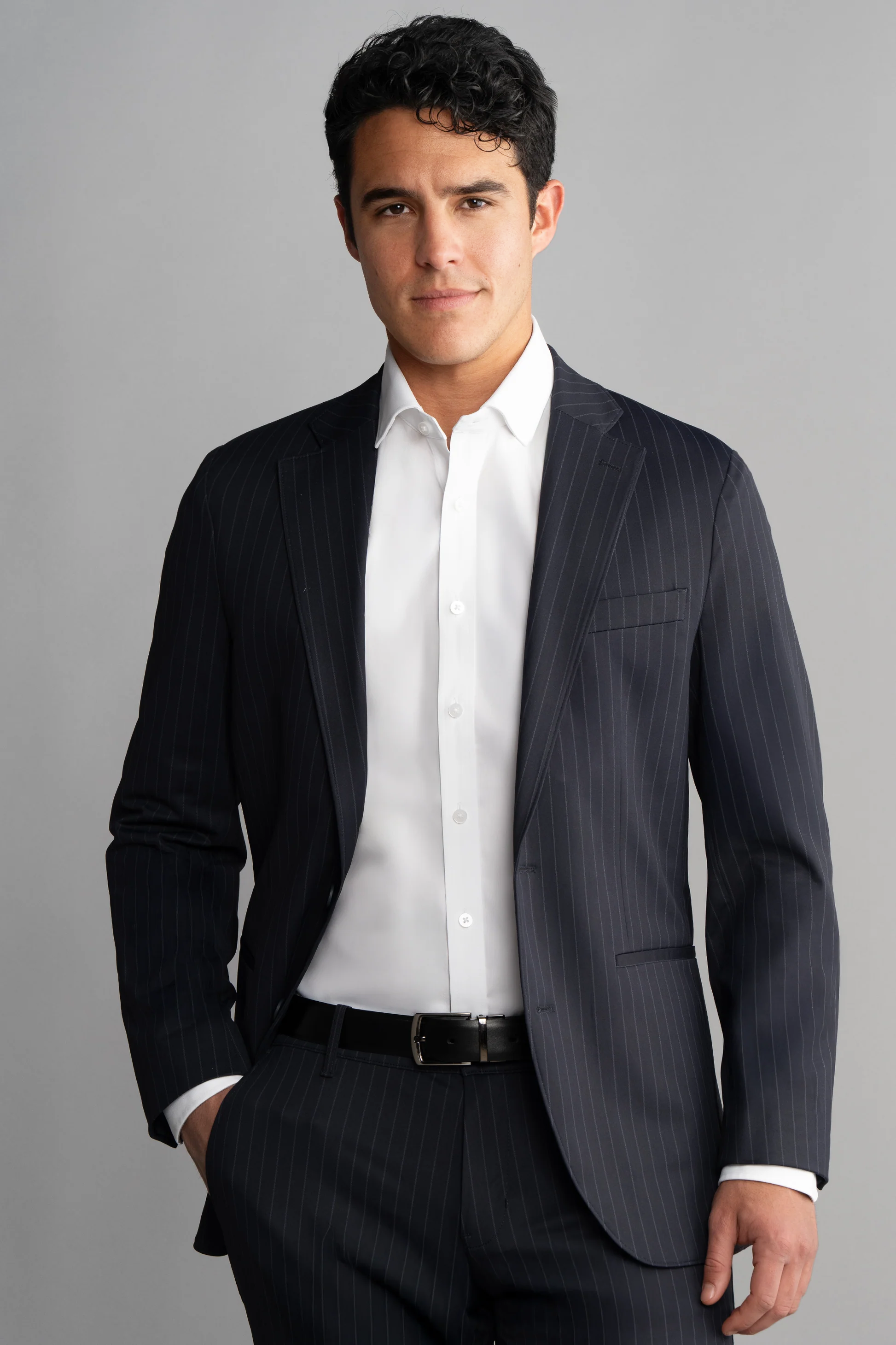 A man with short dark curly hair wears the Twillory Performance Blazer in Navy Stripe over a white dress shirt, unbuttoned at the collar. He stands with one hand in his pocket against a plain gray background, looking confidently at the camera.