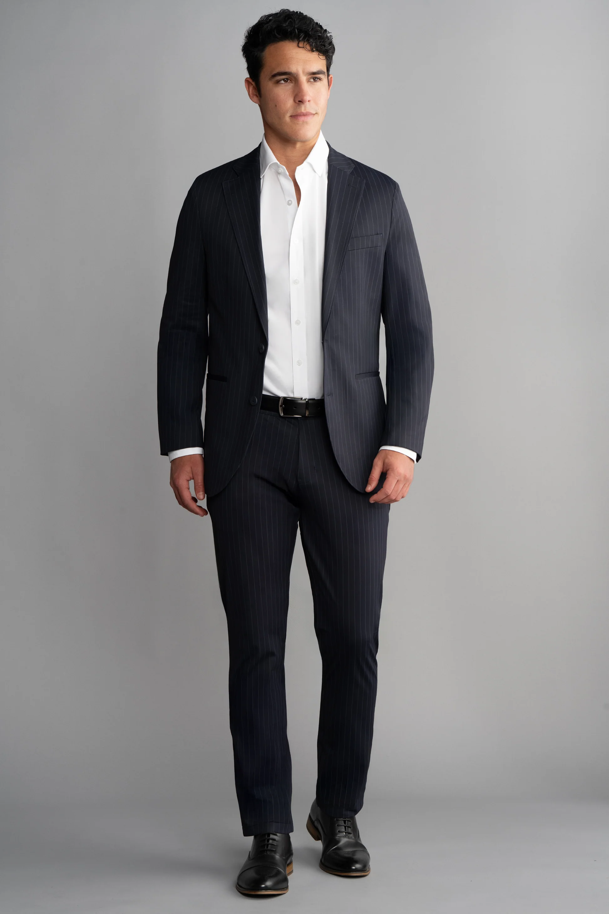 A man with short, dark curly hair and a neutral expression stands against a plain gray background wearing the Twillory Performance Blazer // Navy Stripe over a white dress shirt, styled with a black belt and black dress shoes.