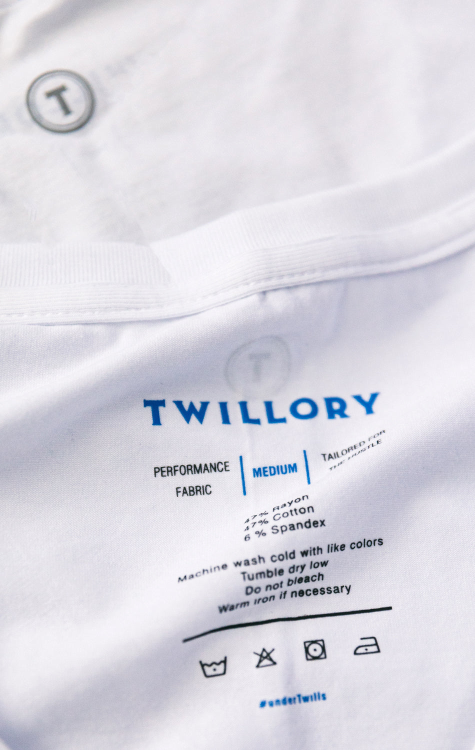 A close-up of a white Twillory Performance Undershirt 3 Pack // Crew Neck shows the inside label with brand name in blue, performance fabric details, medium size, care instructions, and laundry symbols. A button and part of the collar appear at the top.