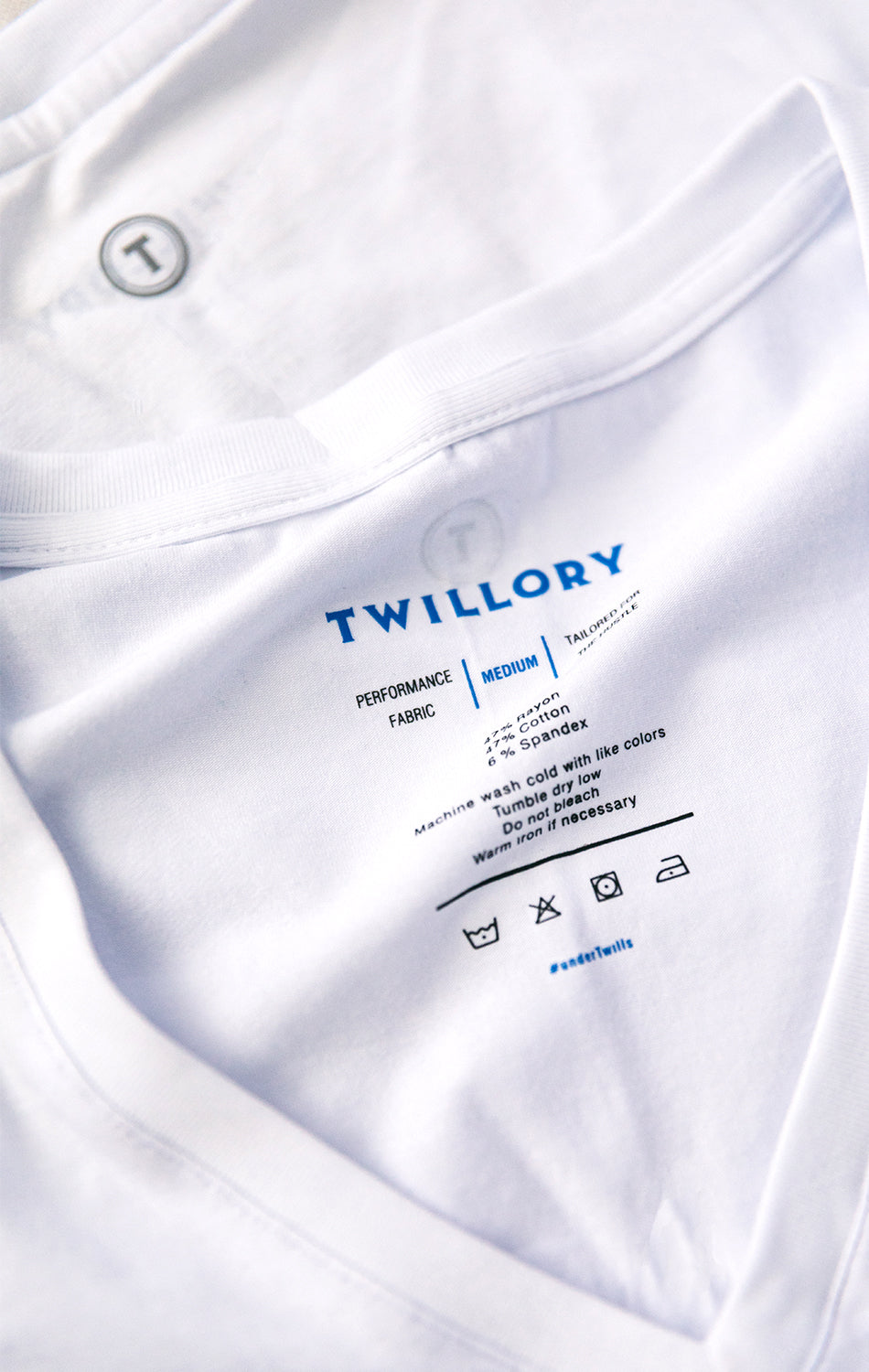 Close-up of a white Twillory Performance Undershirt 3 Pack // V Neck, featuring its inner collar label with blue and black text showing size (medium), care info, fabric details, laundry symbols, #settleforbill; another shirt with "T" logo is beneath.