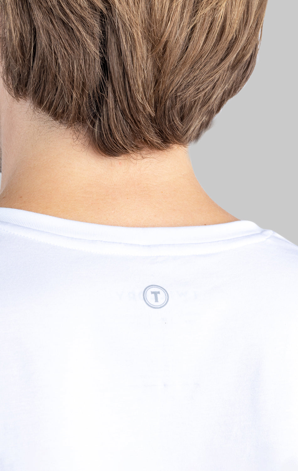 Close-up of the back of a person wearing a white Twillory Performance Undershirt 3 Pack // V Neck. Their short light brown hair partly covers the neckline, revealing a subtle circular “T” logo on the upper back. Gray background.