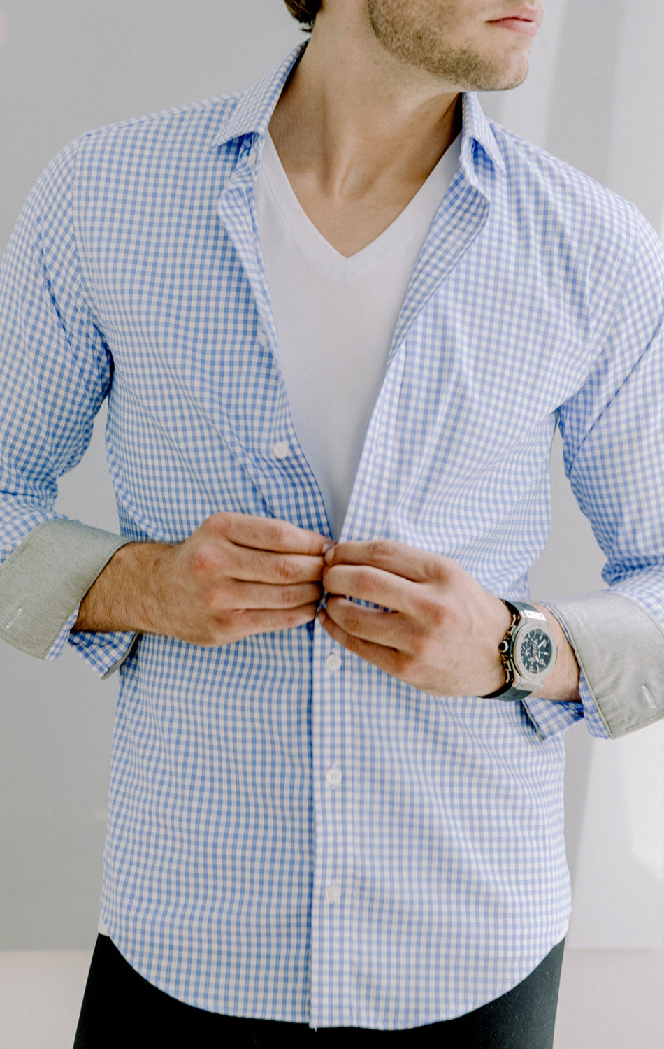 A man with short brown hair and light skin, wearing a Twillory Performance Undershirt 3 Pack // V Neck under a blue checkered shirt, buttons his shirt. His face is partially out of frame; he wears a silver watch. The background is blurred and neutral.