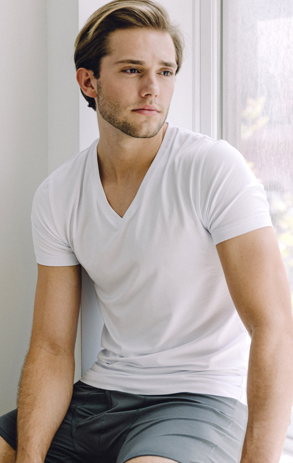A young man with light brown hair relaxes by a sunlit window, wearing a fitted Twillory Performance Undershirt 3 Pack // V Neck and gray shorts, creating a calm, relaxed atmosphere.