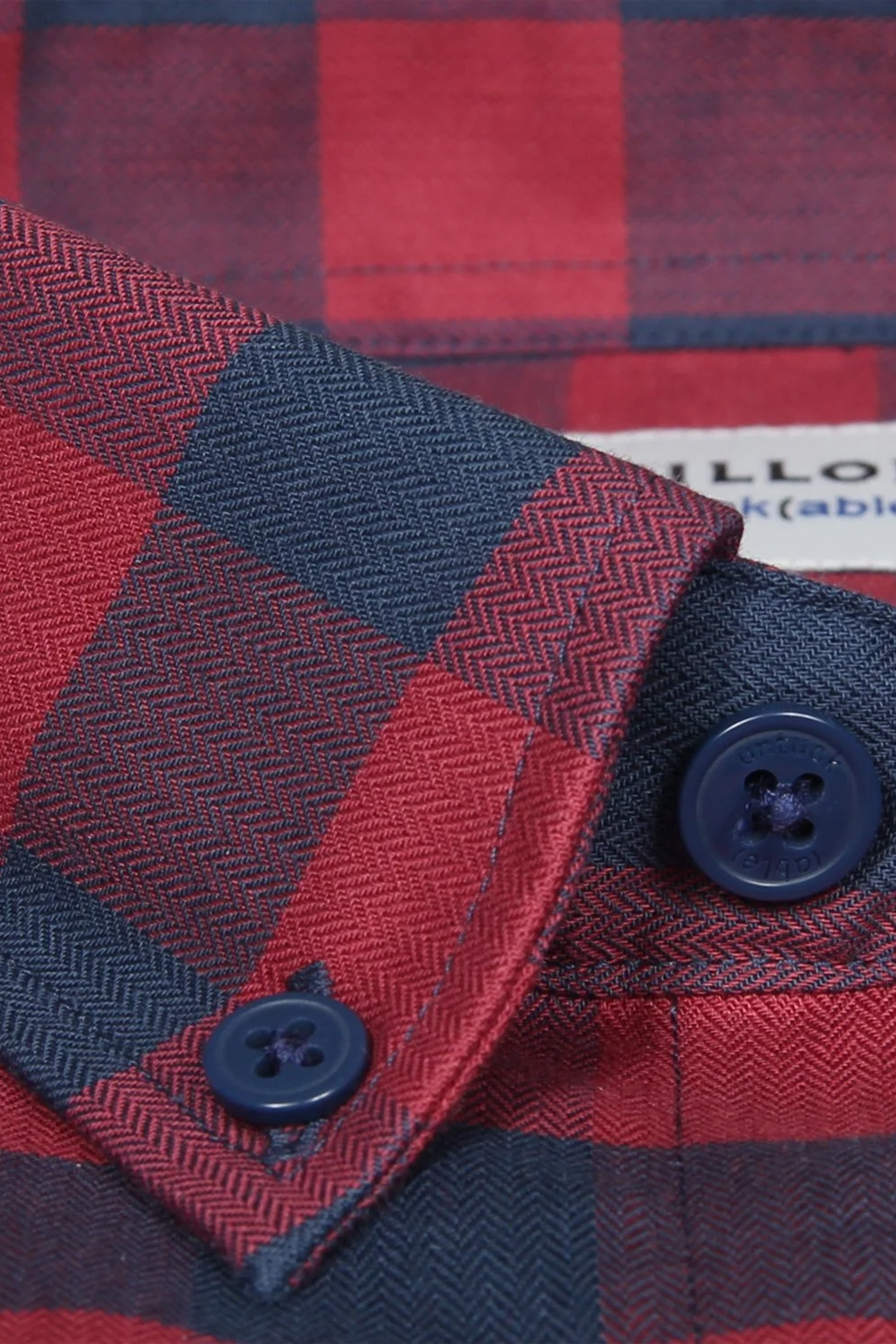 Close-up of Twillory's untuck(able) Barnyard Gingham shirt cuff in red and navy plaid herringbone, made from non-iron stretch fabric. The smart casual cuff has two dark blue buttons, with part of the label visible in the background.