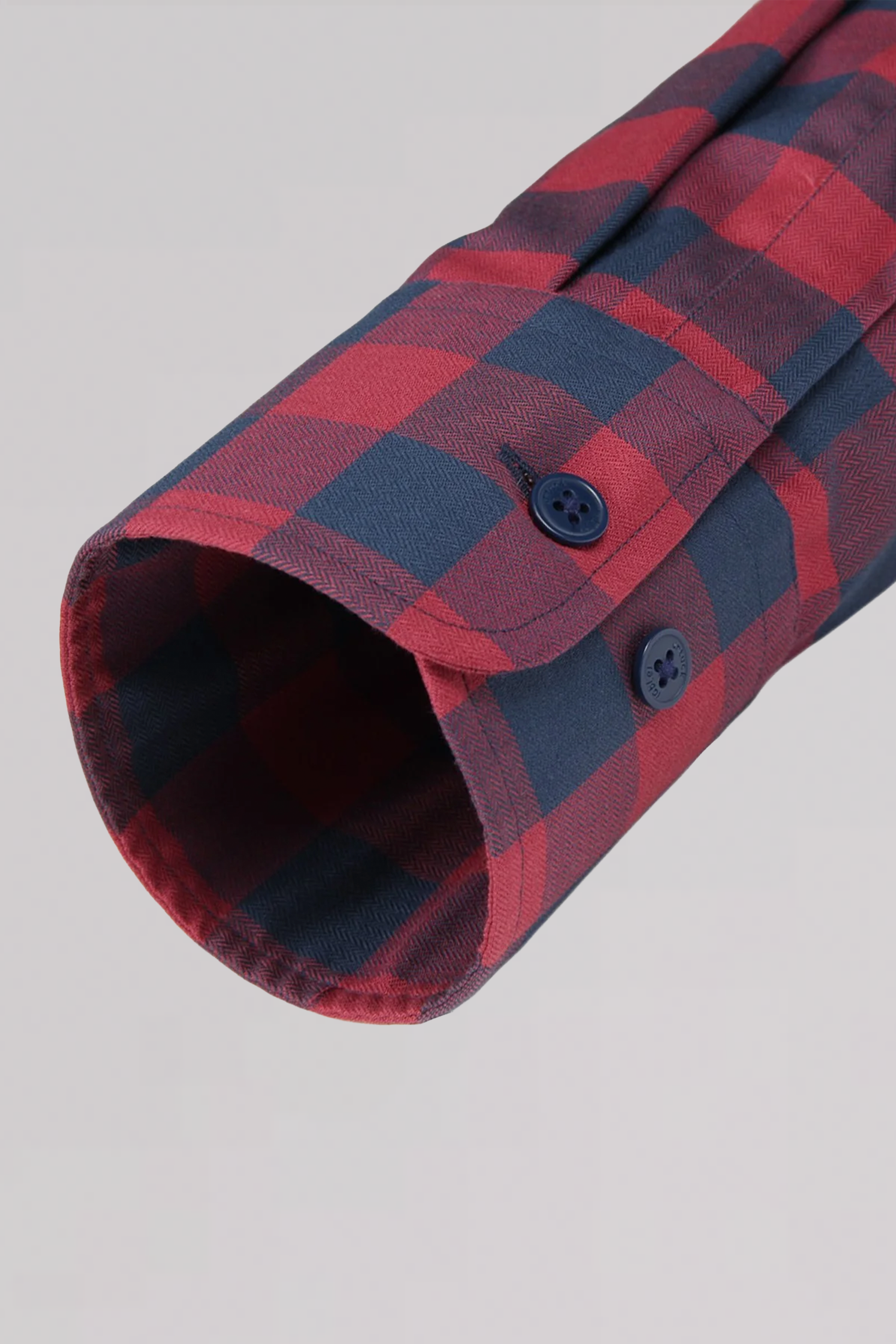 A close-up of the Twillory untuck(able) Barnyard Gingham shirt sleeve in red and navy plaid non-iron stretch fabric, with a cuff featuring two dark blue buttons, set against a plain light gray background.
