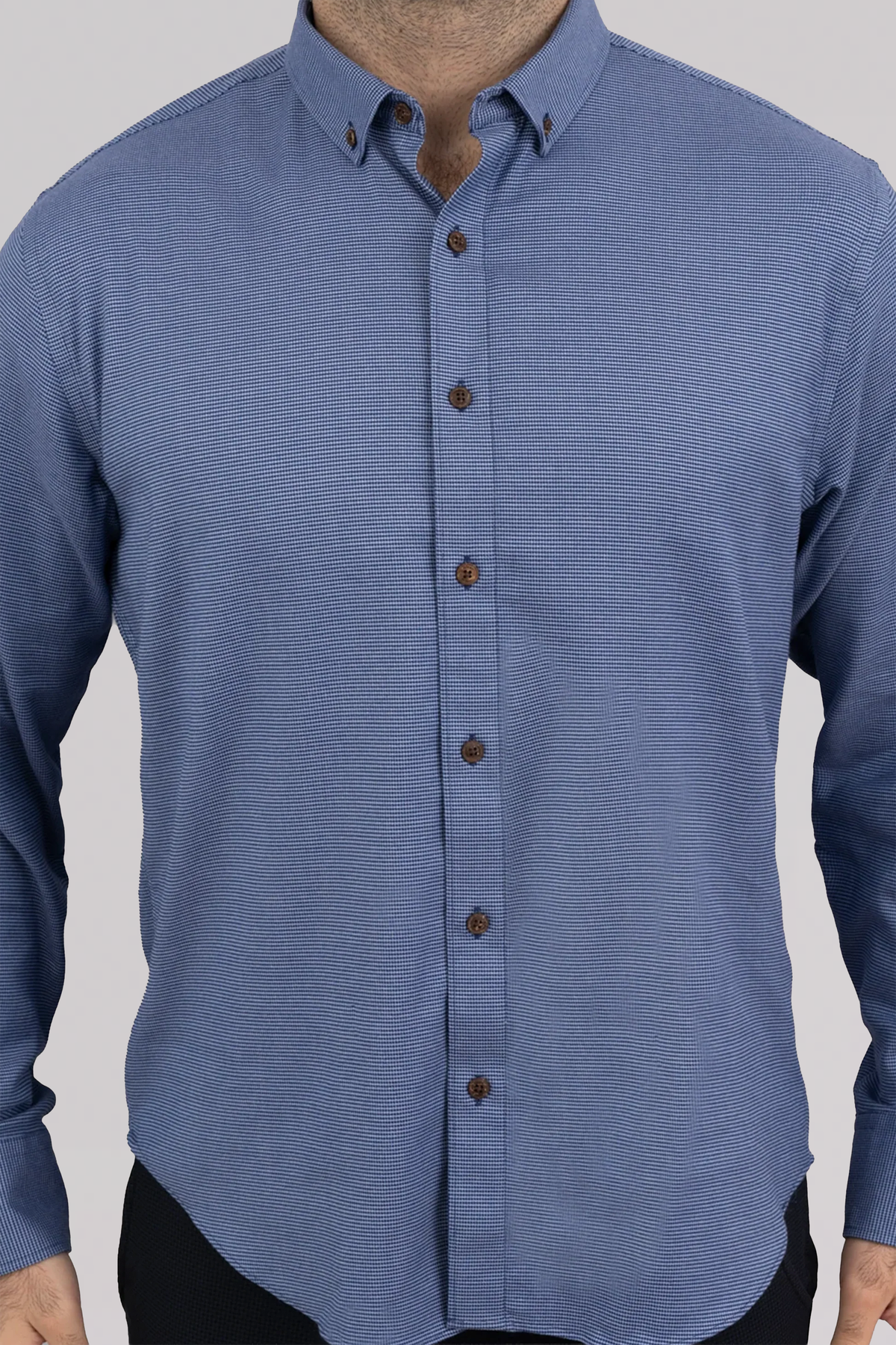 A man wears the Twillory untuck(able) Blue Puppytooth shirt—a long-sleeved, light blue shirt with a subtle puppytooth pattern, classic collar with button points, and brown front buttons. Image cropped at neck and mid-thigh.